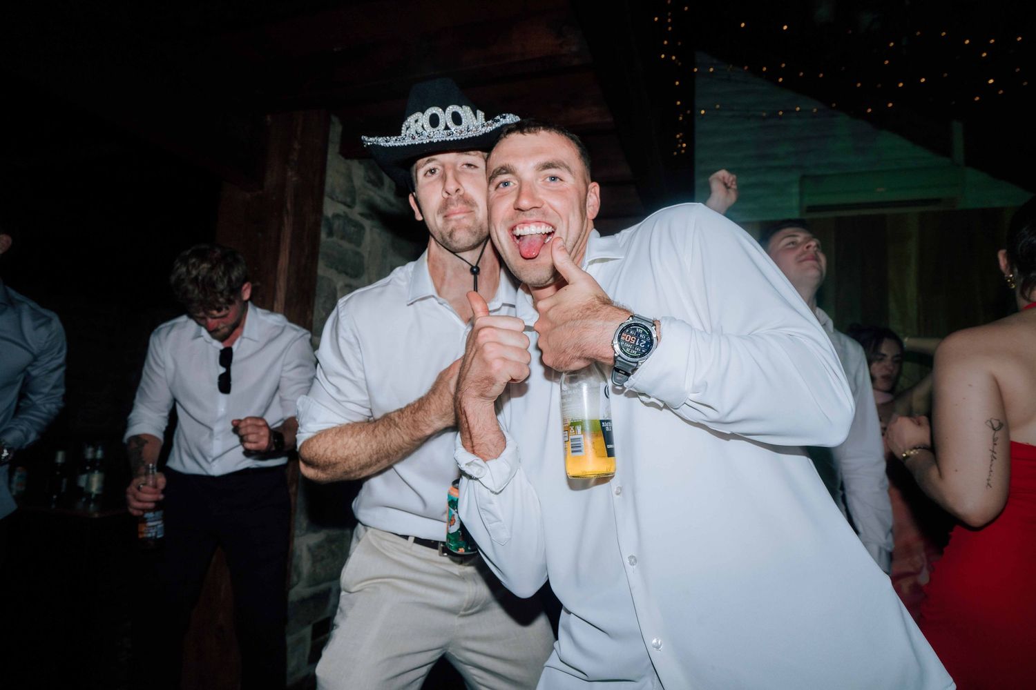 Groom wearing cowboy hat posing with friend giving thumbs up at wedding reception.