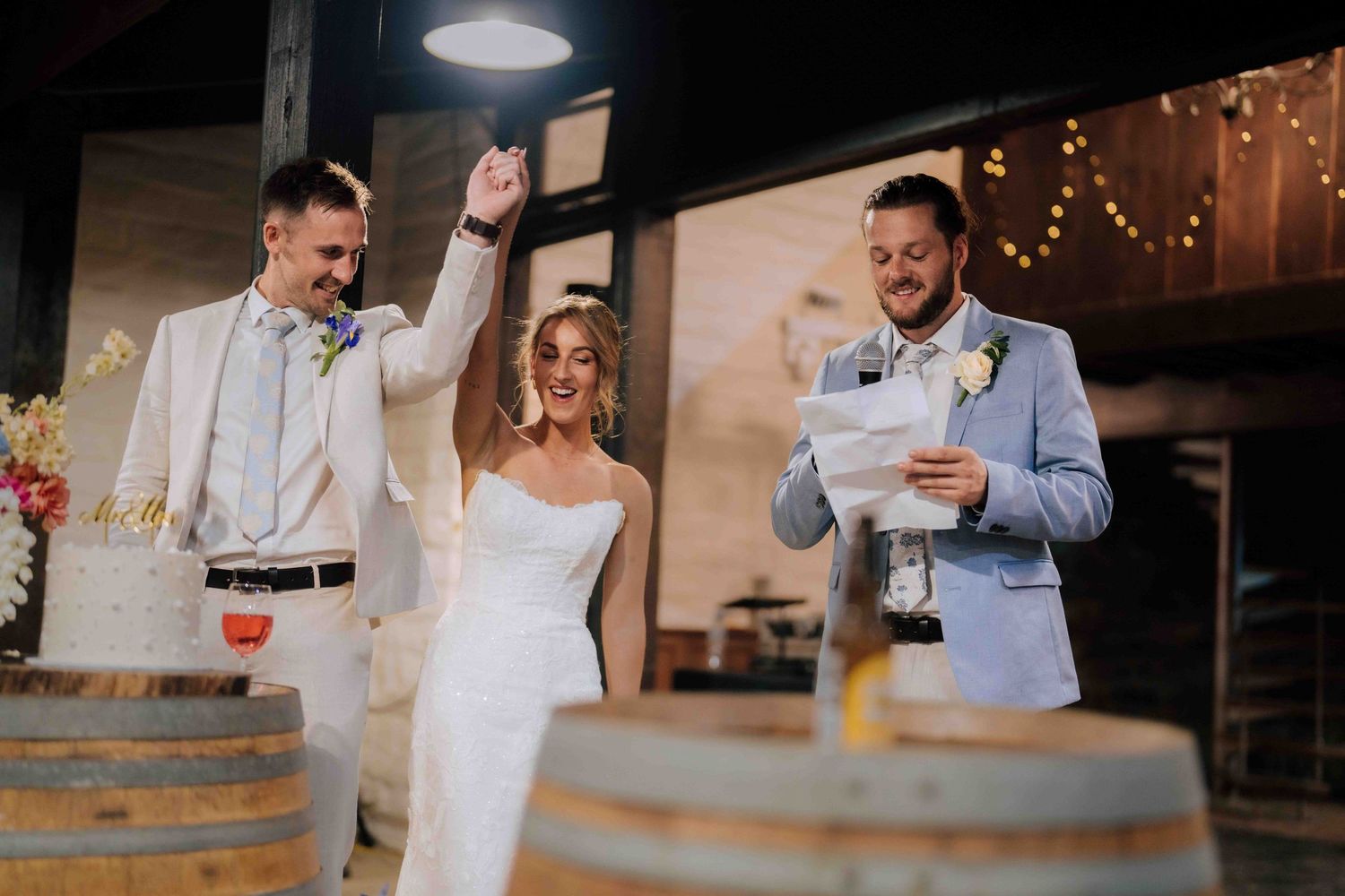 Bride and groom celebrating with officiant beside wedding cake on barrel at rustic venue.
