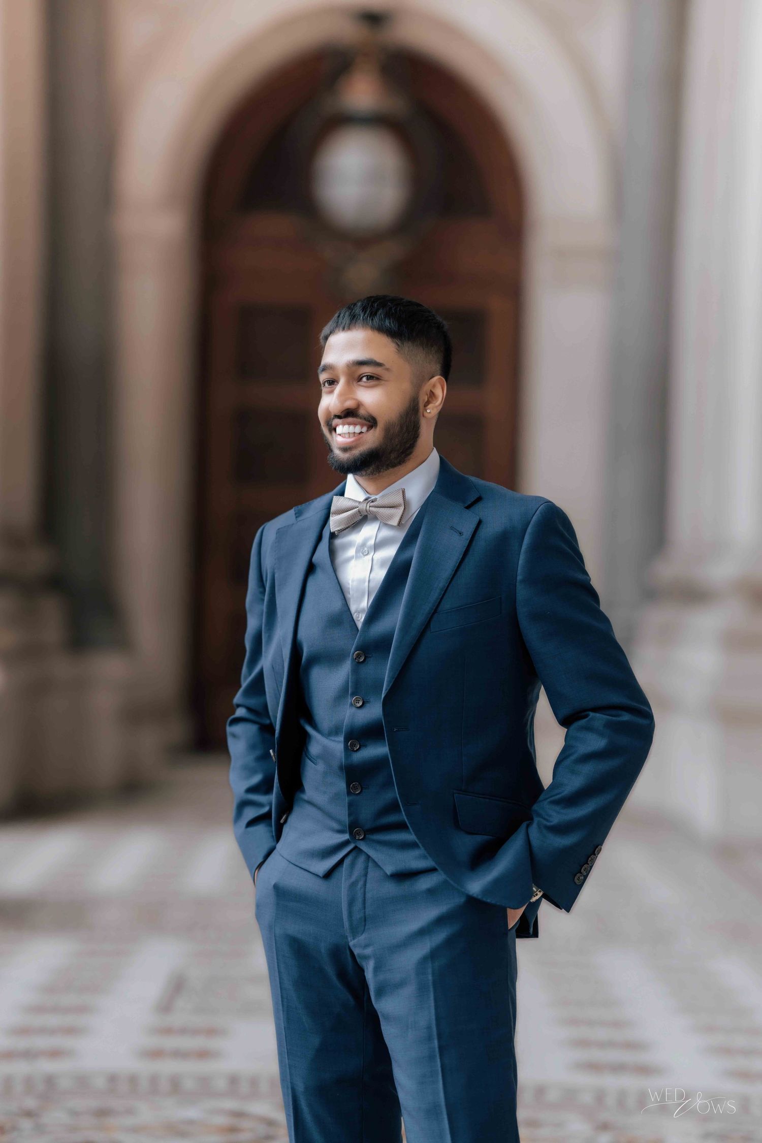 Handsome groom smiling in navy three-piece suit with bow tie in elegant arched hallway.
