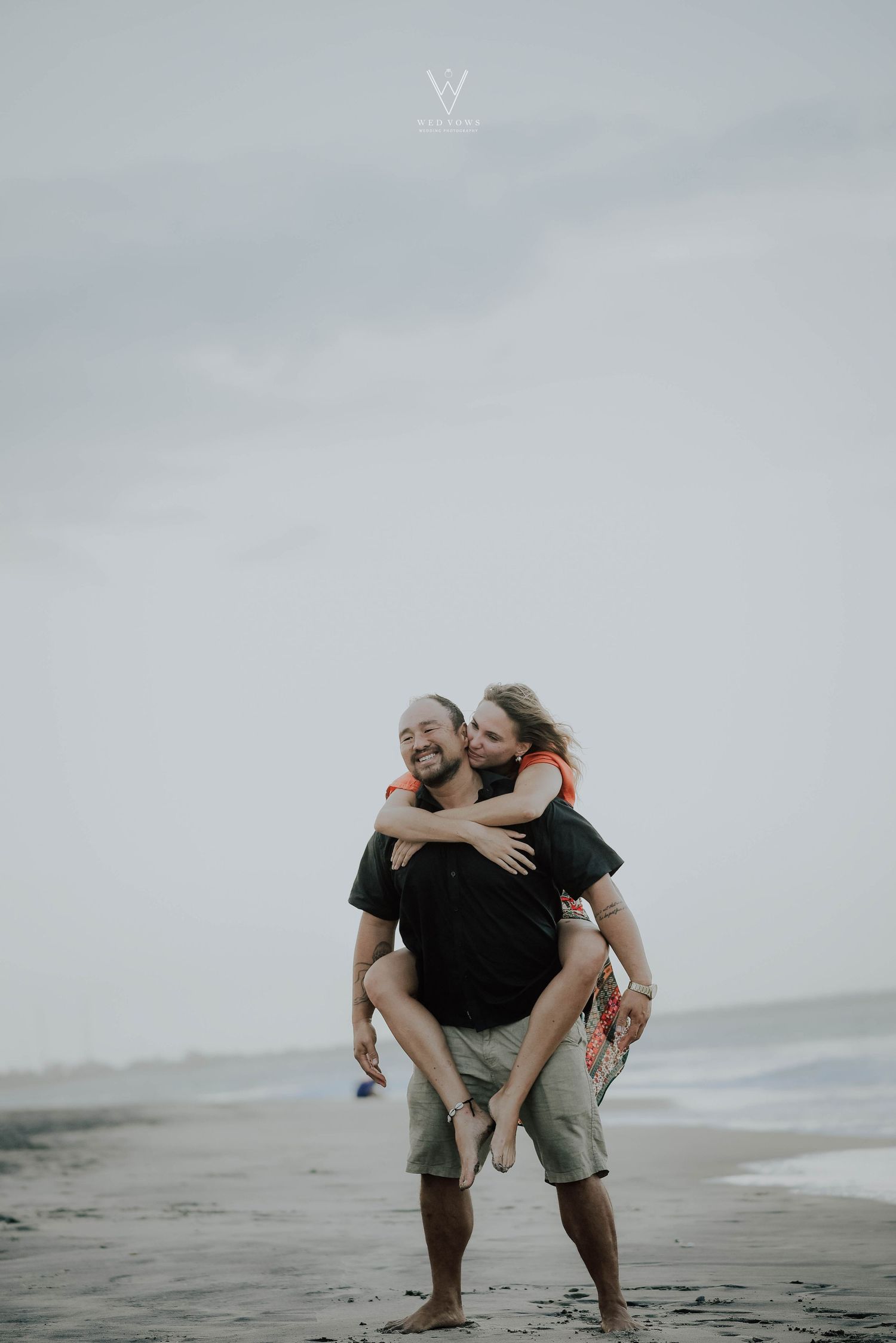 Couple sharing a joyful piggyback ride on a beach, man smiling as woman hugs him from behind.