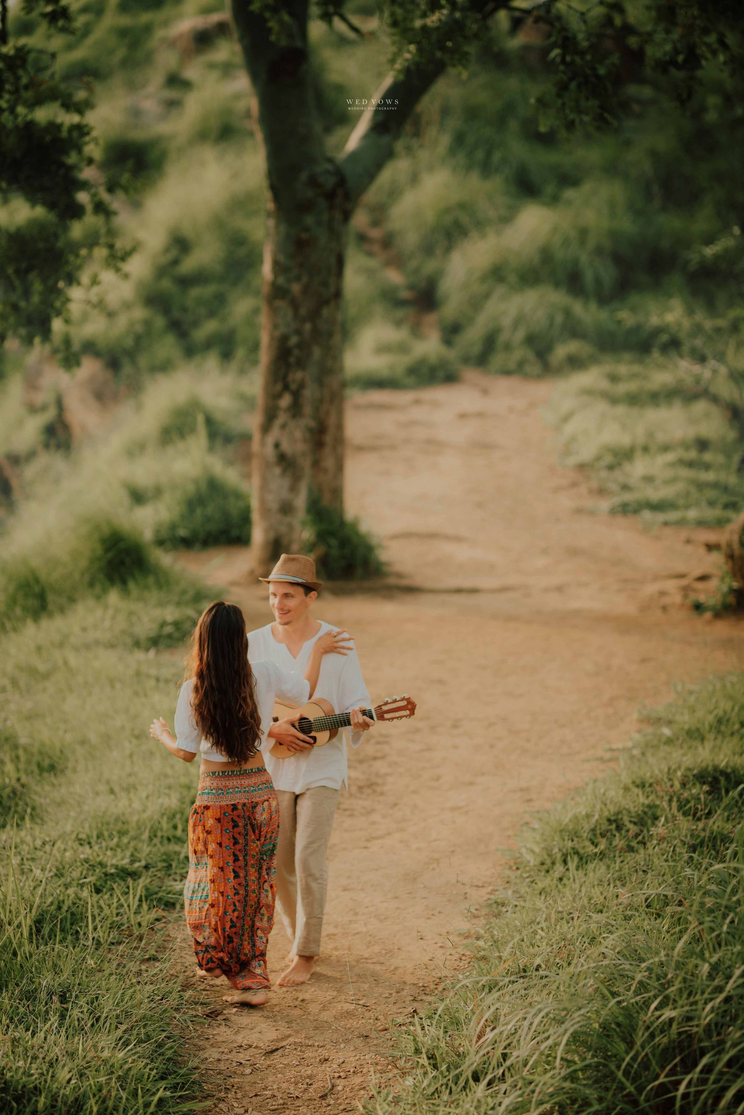 Man playing ukulele serenades woman on scenic dirt trail surrounded by greenery.