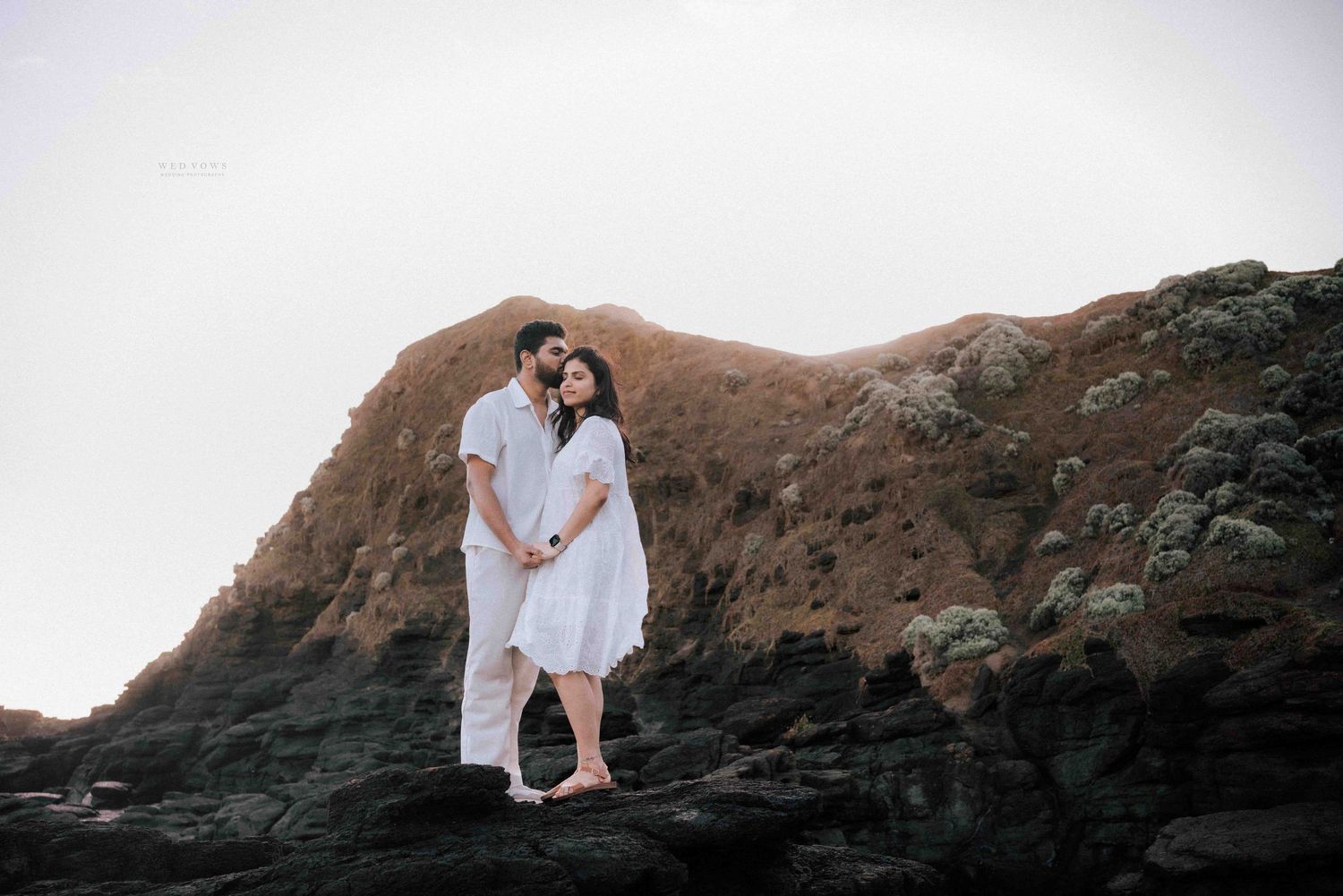 Couple dressed in white embracing on dark volcanic rocks with golden hillside backdrop.