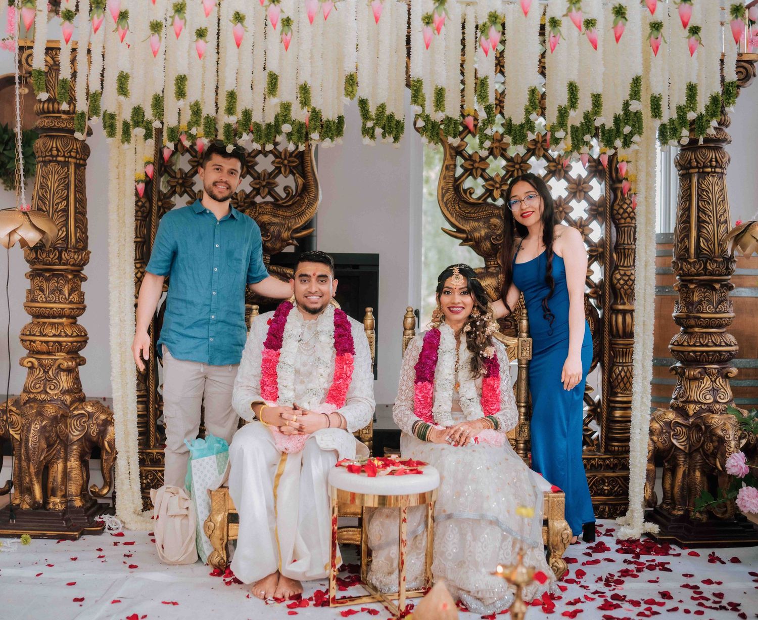 Bride and groom seated at ornate mandap with two guests standing behind during Indian wedding ceremony.