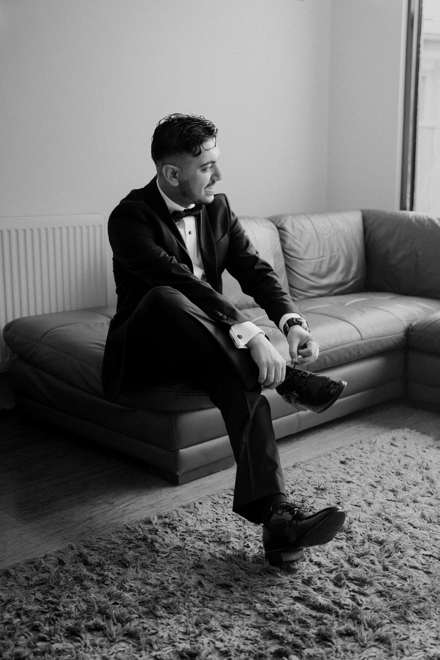 Groom in tuxedo sitting on leather sofa adjusting cufflinks before wedding ceremony in black and white.