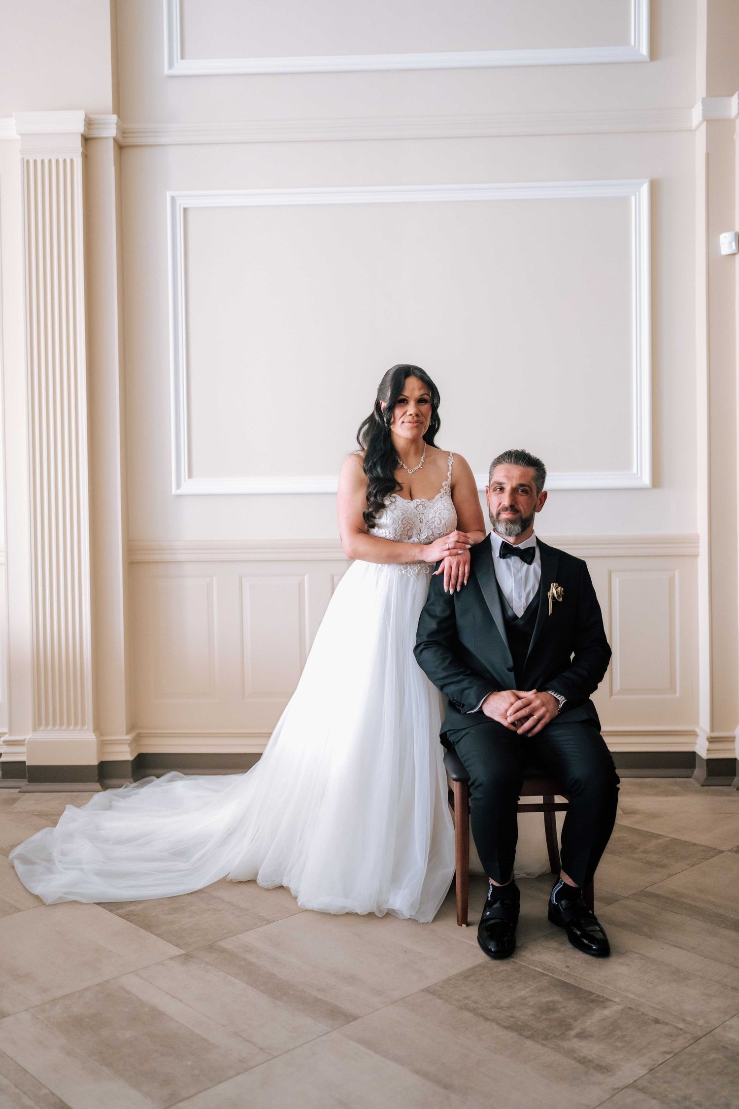 Bride and groom posing elegantly in a bright white room, groom seated in tuxedo, bride standing in white ball gown.