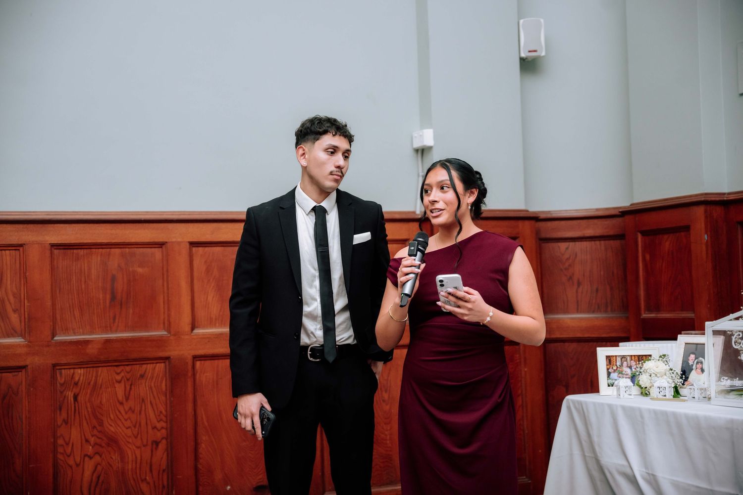 Man and woman in formal attire giving a speech at wedding reception holding microphone and phone.