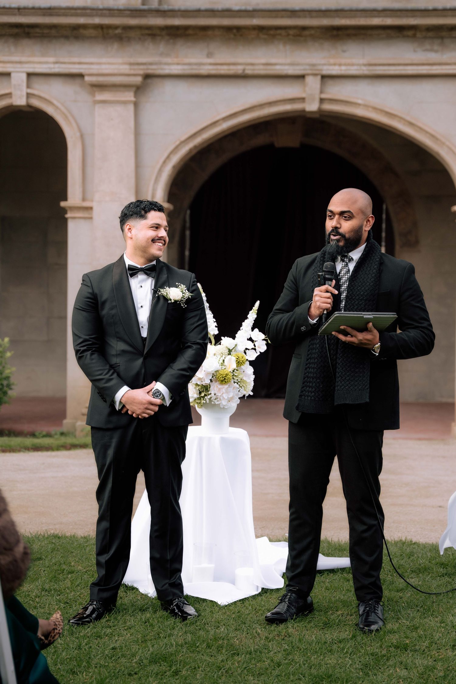 Groom smiling at outdoor wedding ceremony while officiant speaks into microphone beside floral arrangement.