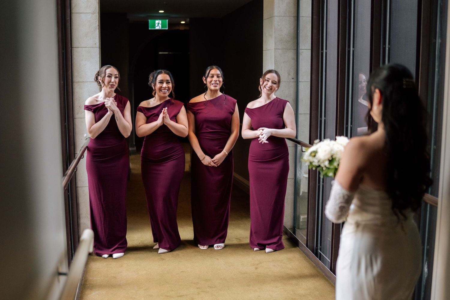 Four bridesmaids in burgundy gowns reacting with joy as bride reveals her wedding dress in hallway.