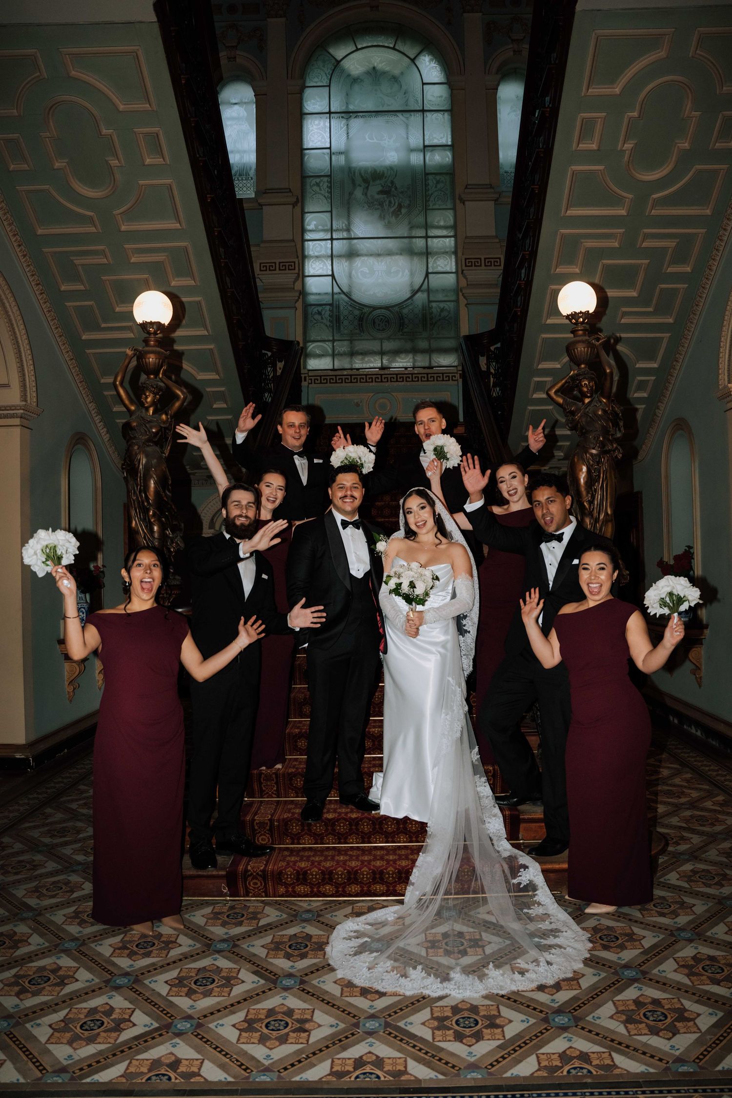 Wedding party poses on ornate staircase with bride in white gown and bridesmaids in burgundy dresses.