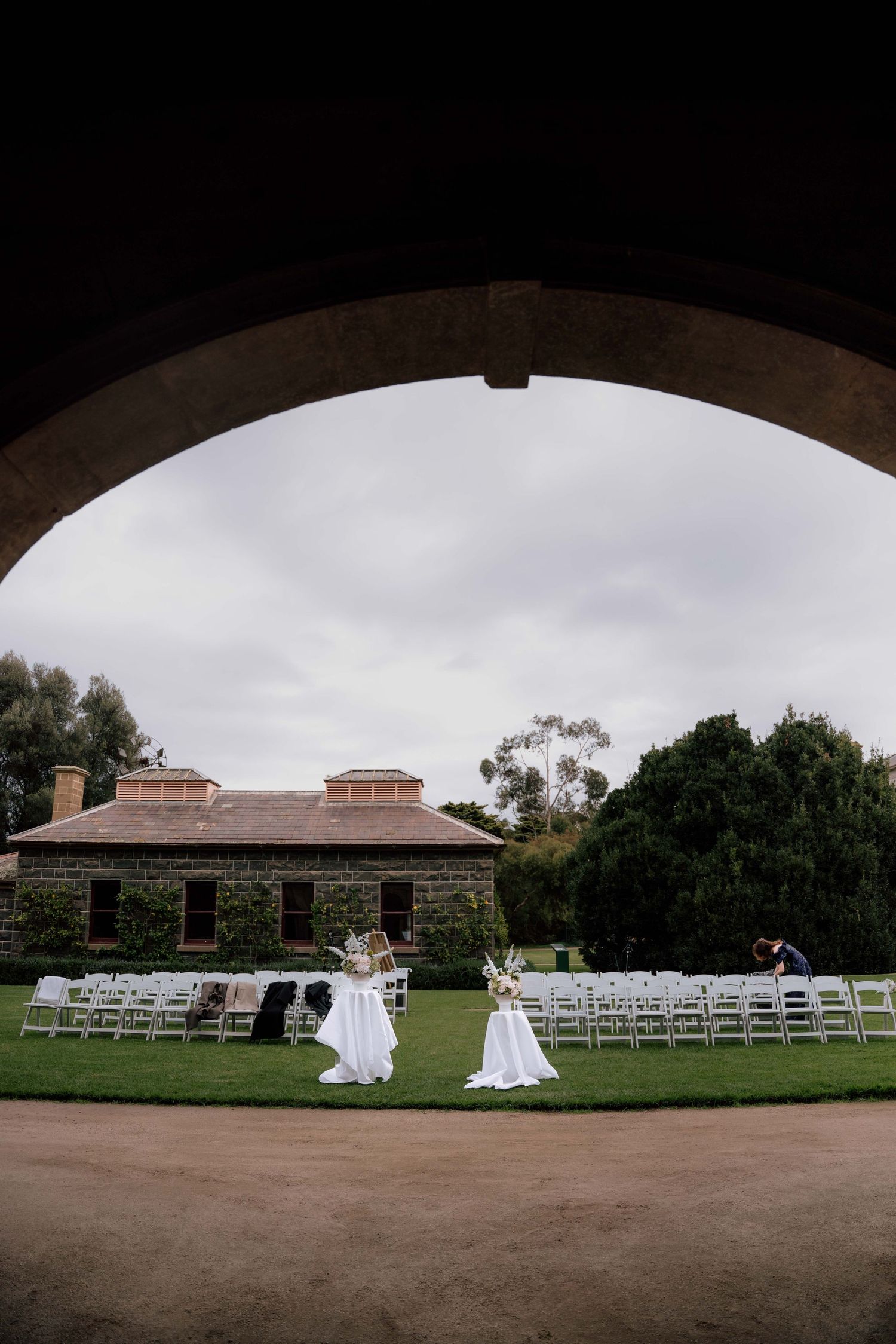 Outdoor wedding ceremony setup with white chairs on green lawn framed by stone arch.