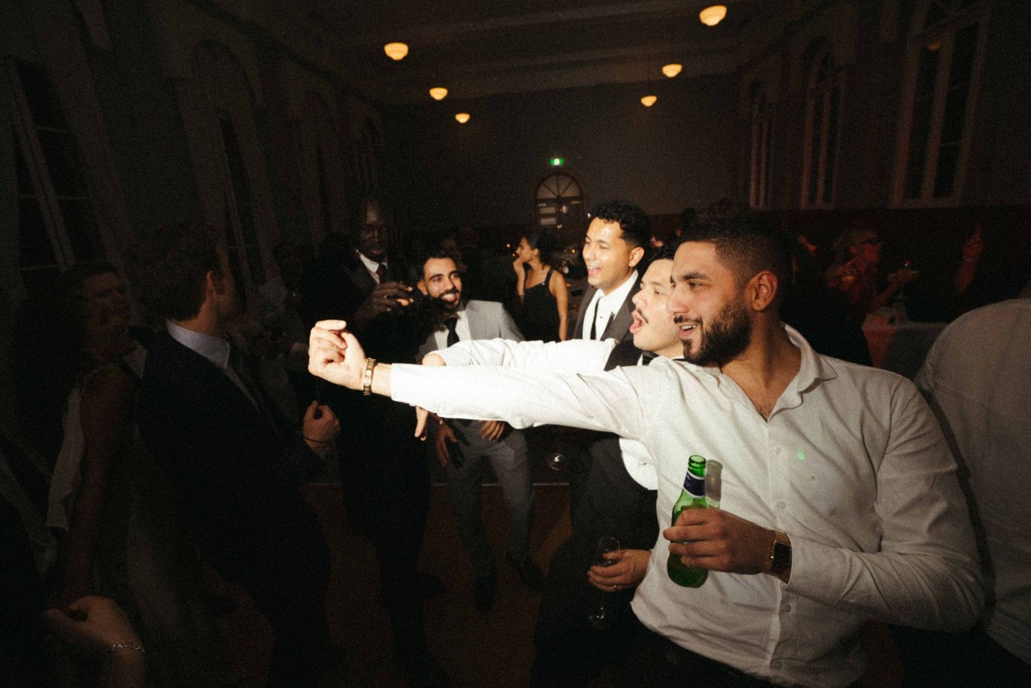 Wedding guest taking selfie while holding beer bottle on crowded reception dance floor at night.