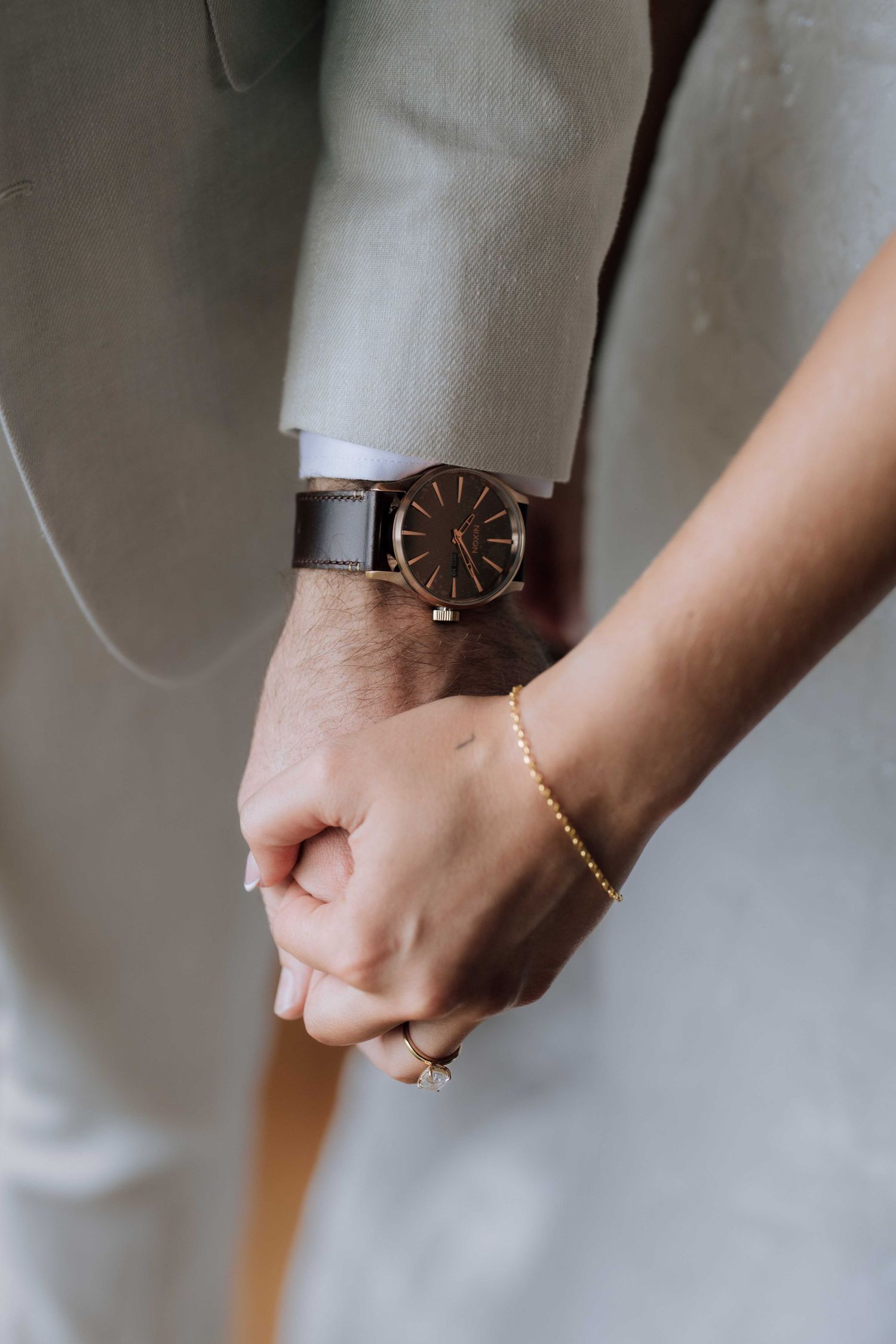 Close-up of bride and groom holding hands showing wooden watch and gold chain bracelet with diamond ring.
