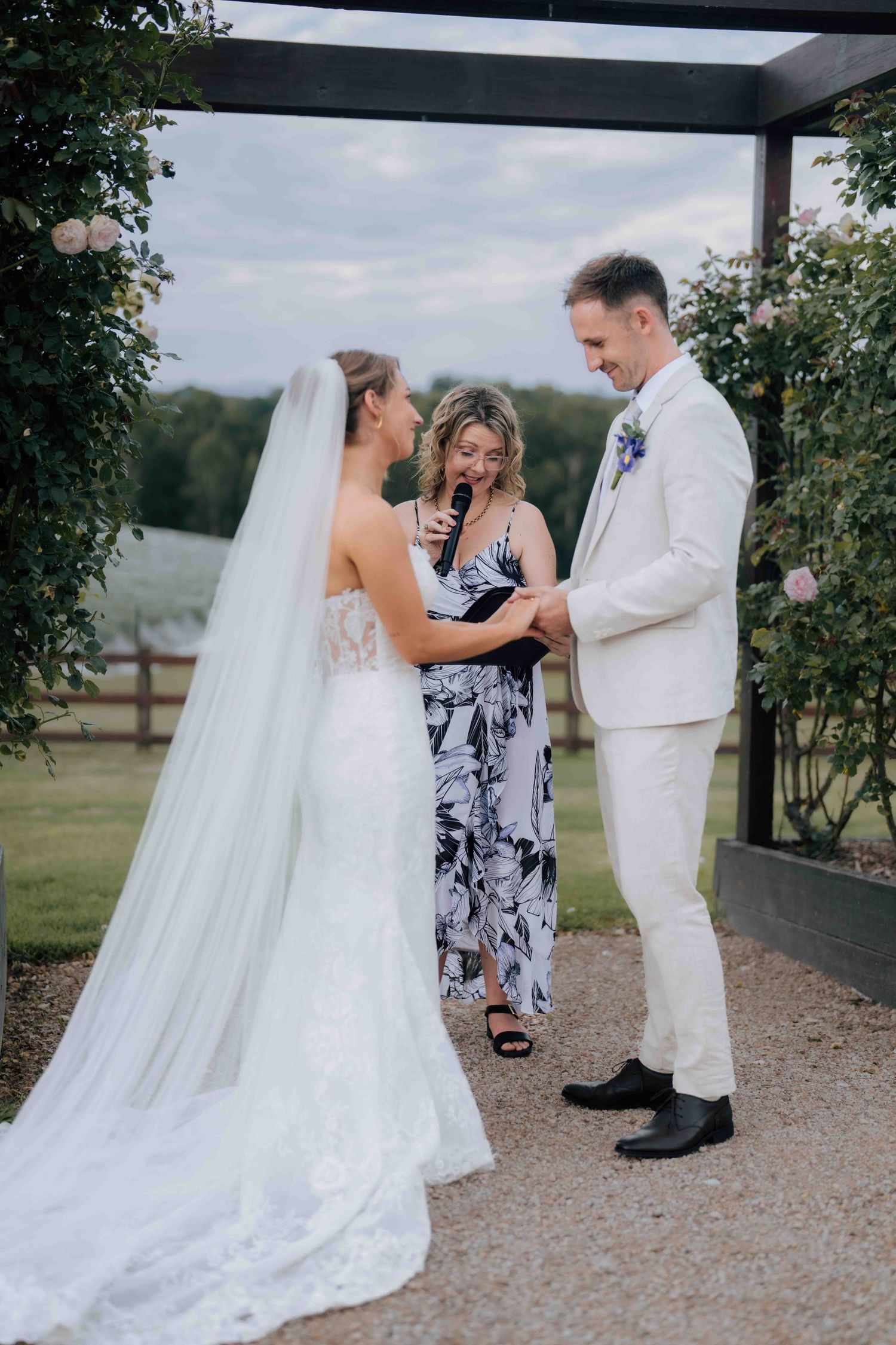 Celebrant reads vows as smiling groom holds brides hands at outdoor rose-adorned wedding ceremony.