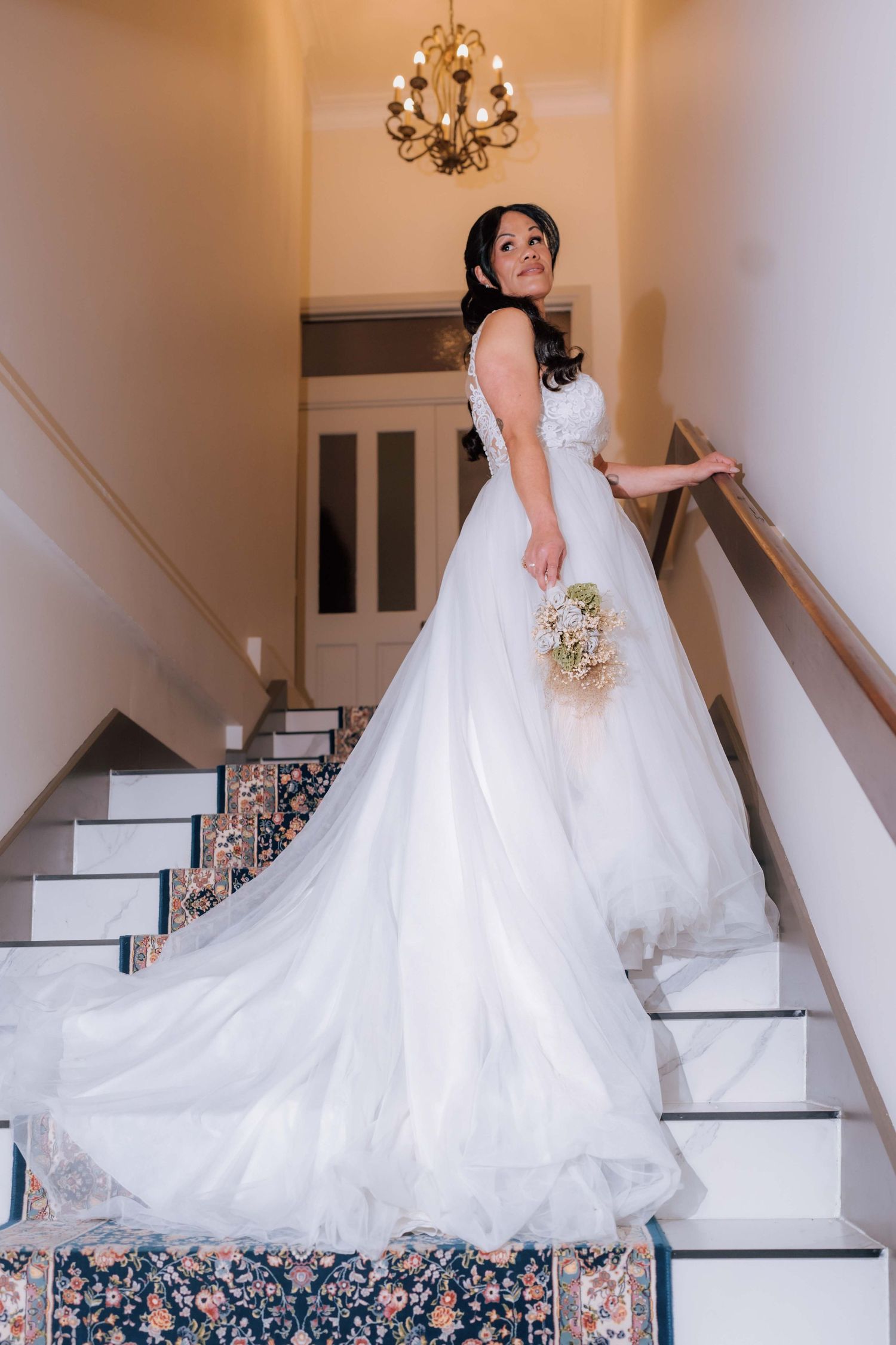 A bride in a long white wedding gown descends a carpeted staircase holding a delicate floral bouquet.