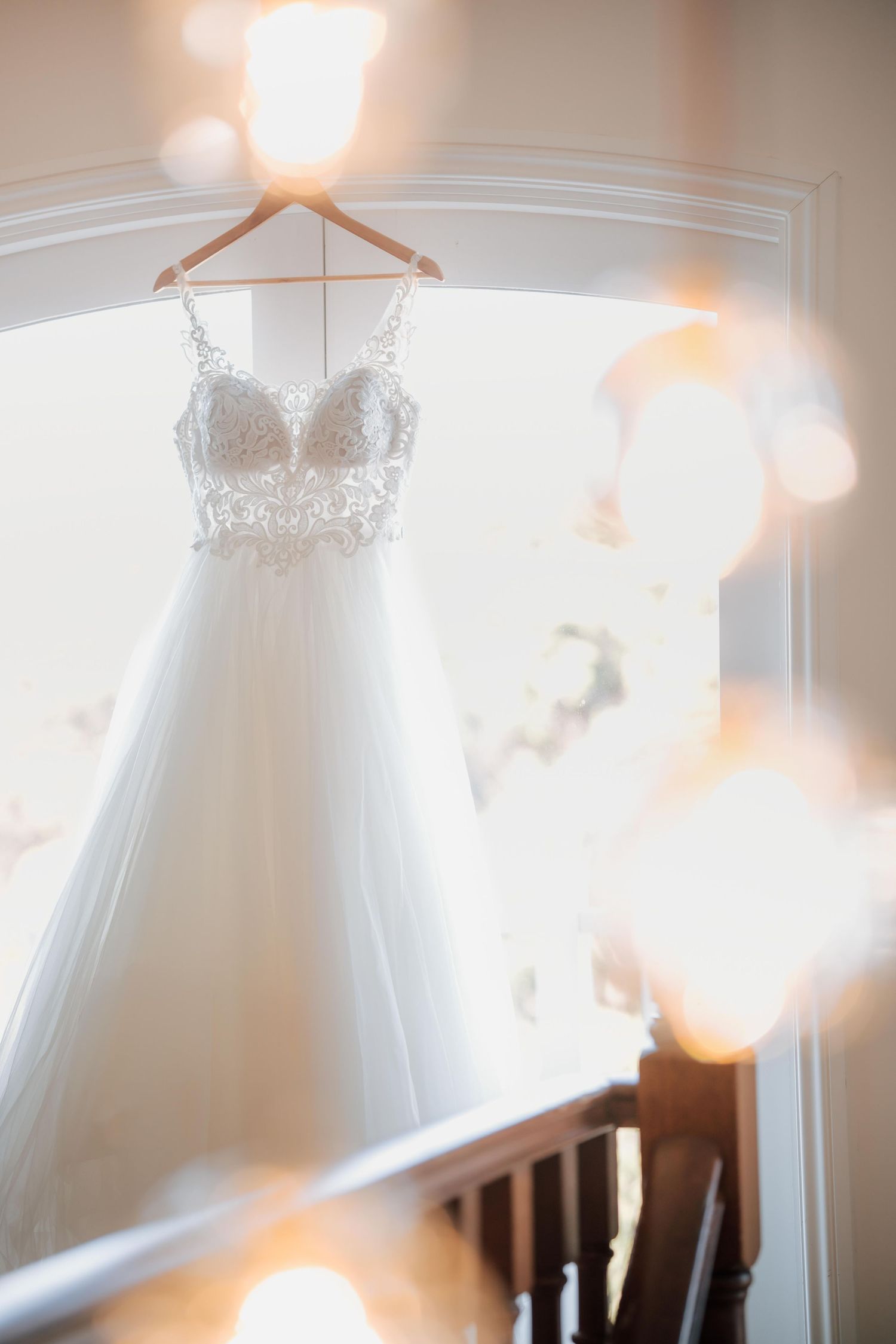 Elegant lace-bodice wedding dress hanging on wooden hanger in a softly lit window with bokeh.