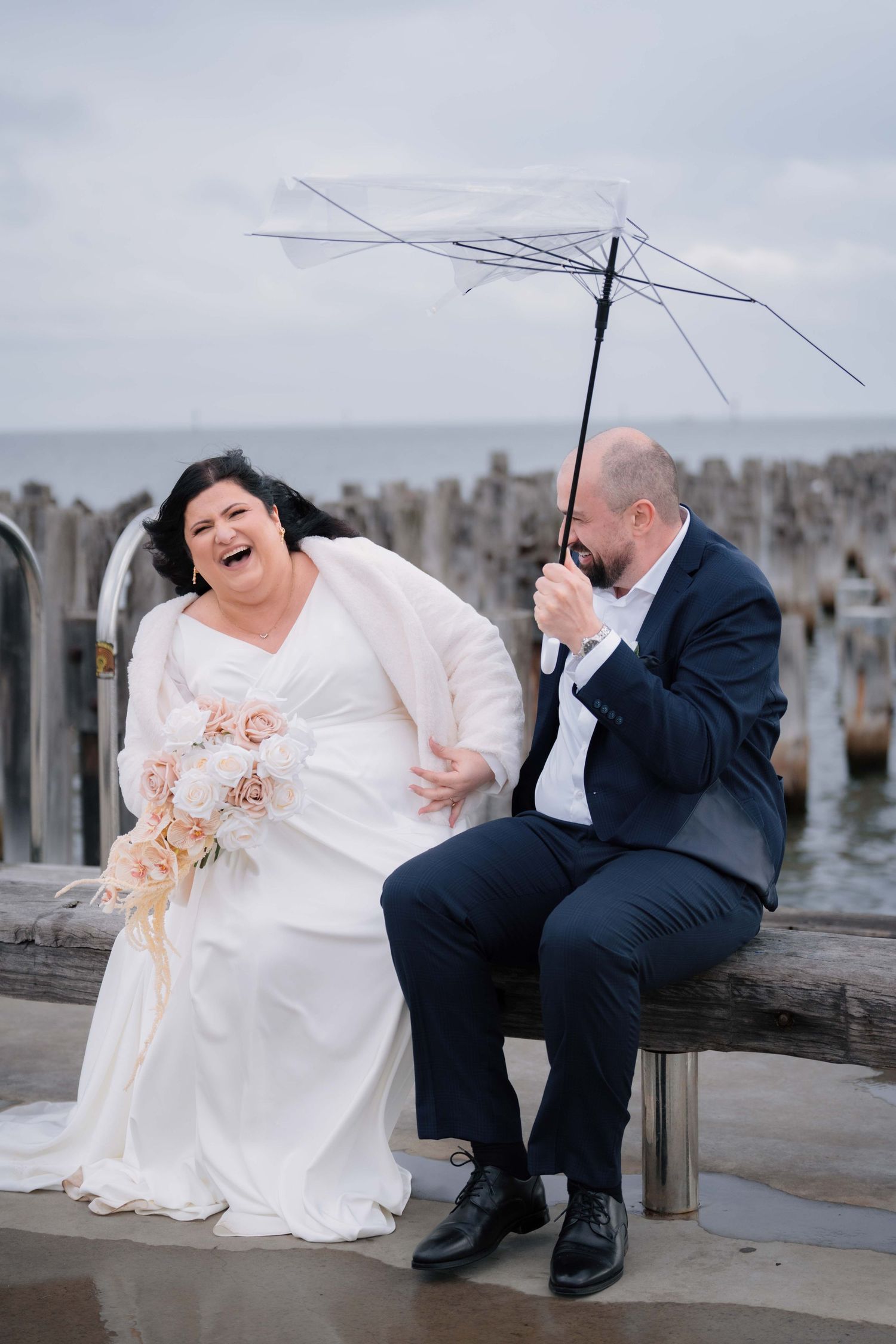 Laughing bride and groom sit on a pier bench with a clear umbrella on a rainy wedding day.