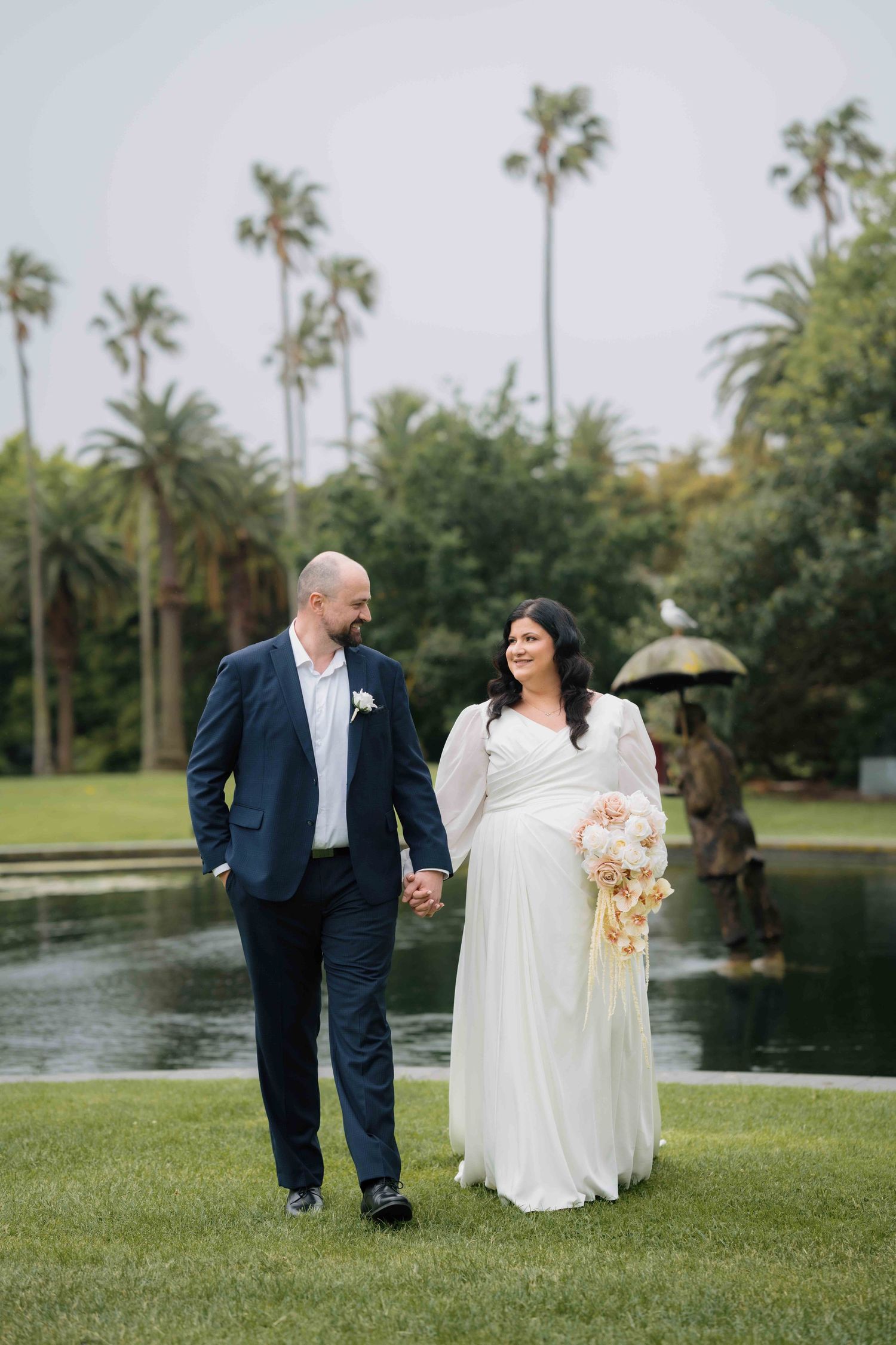 Bride and groom walk hand in hand on green lawn with palm trees and pond in background.