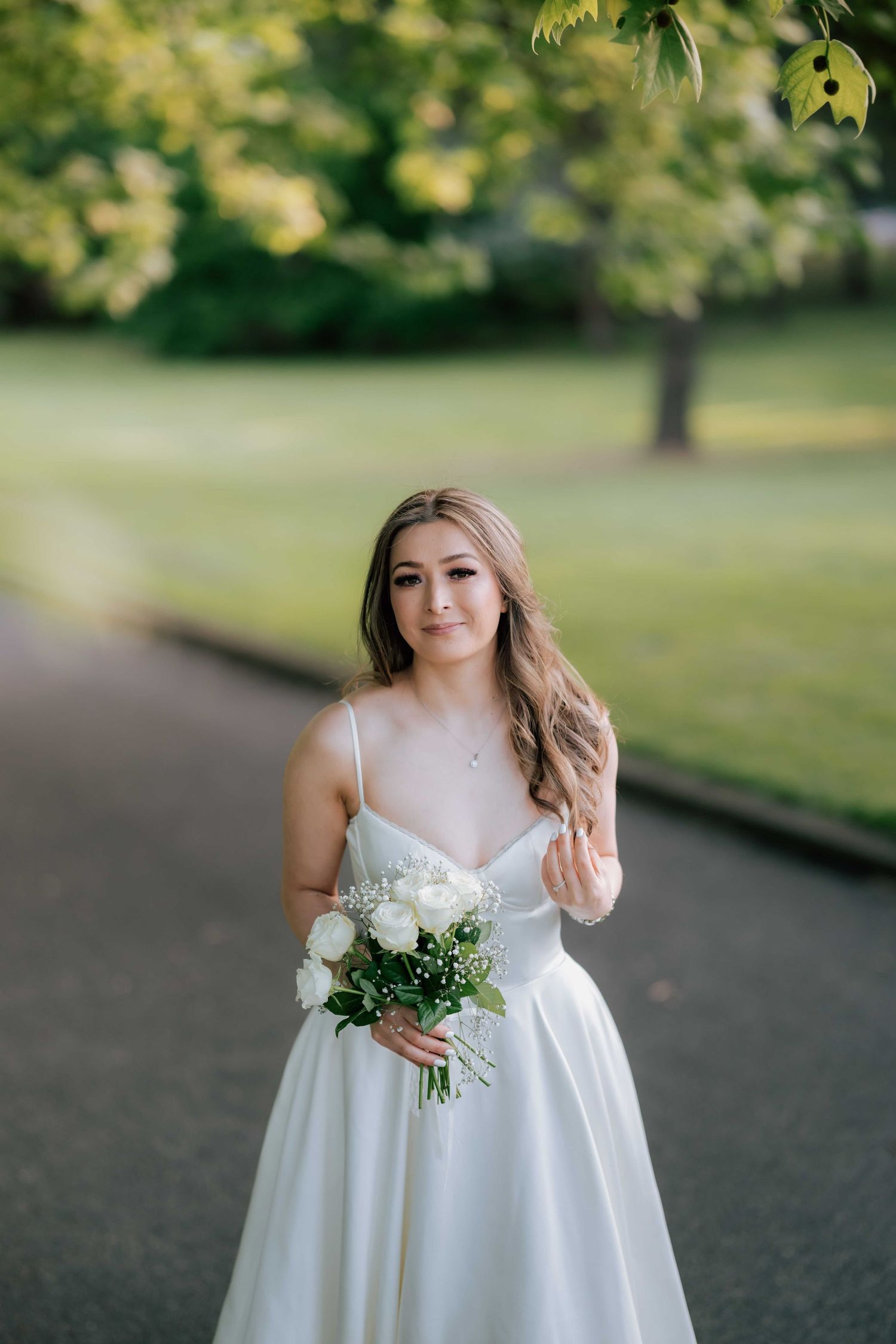Bride in a white satin gown holding white roses while posing on a park path with green trees.