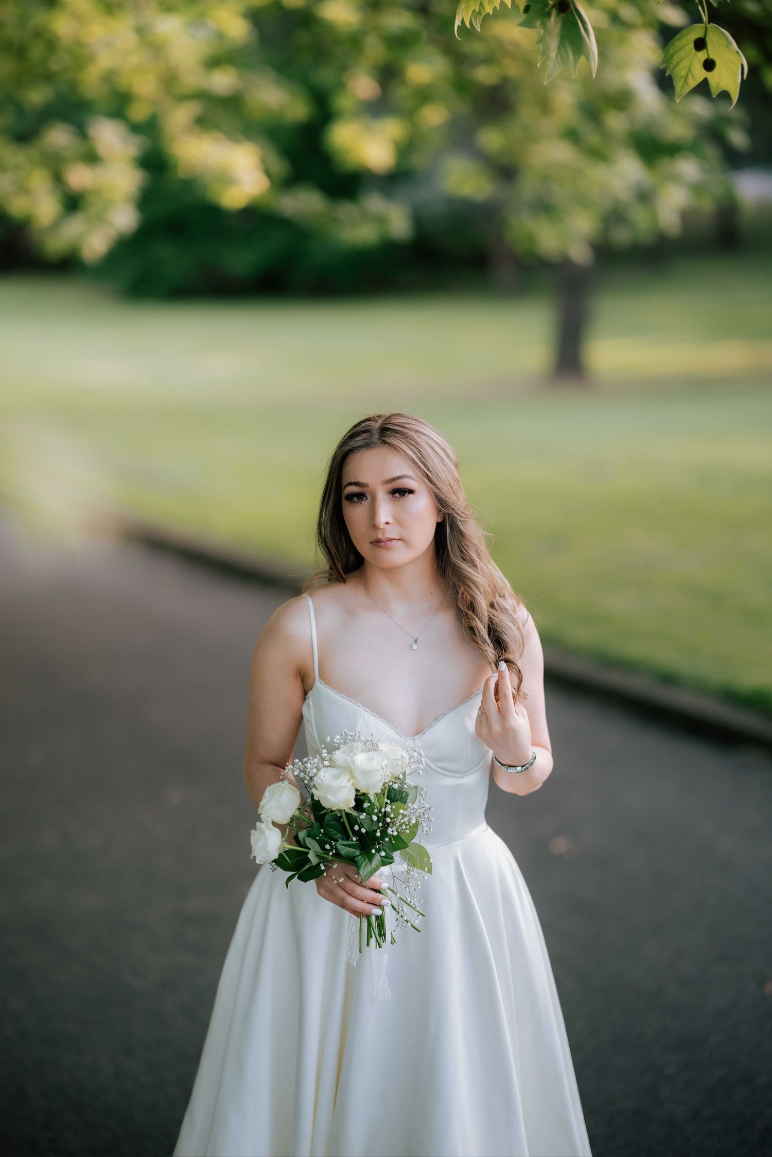 Elegant bride in satin white dress holding white roses bouquet standing in sunlit green park.