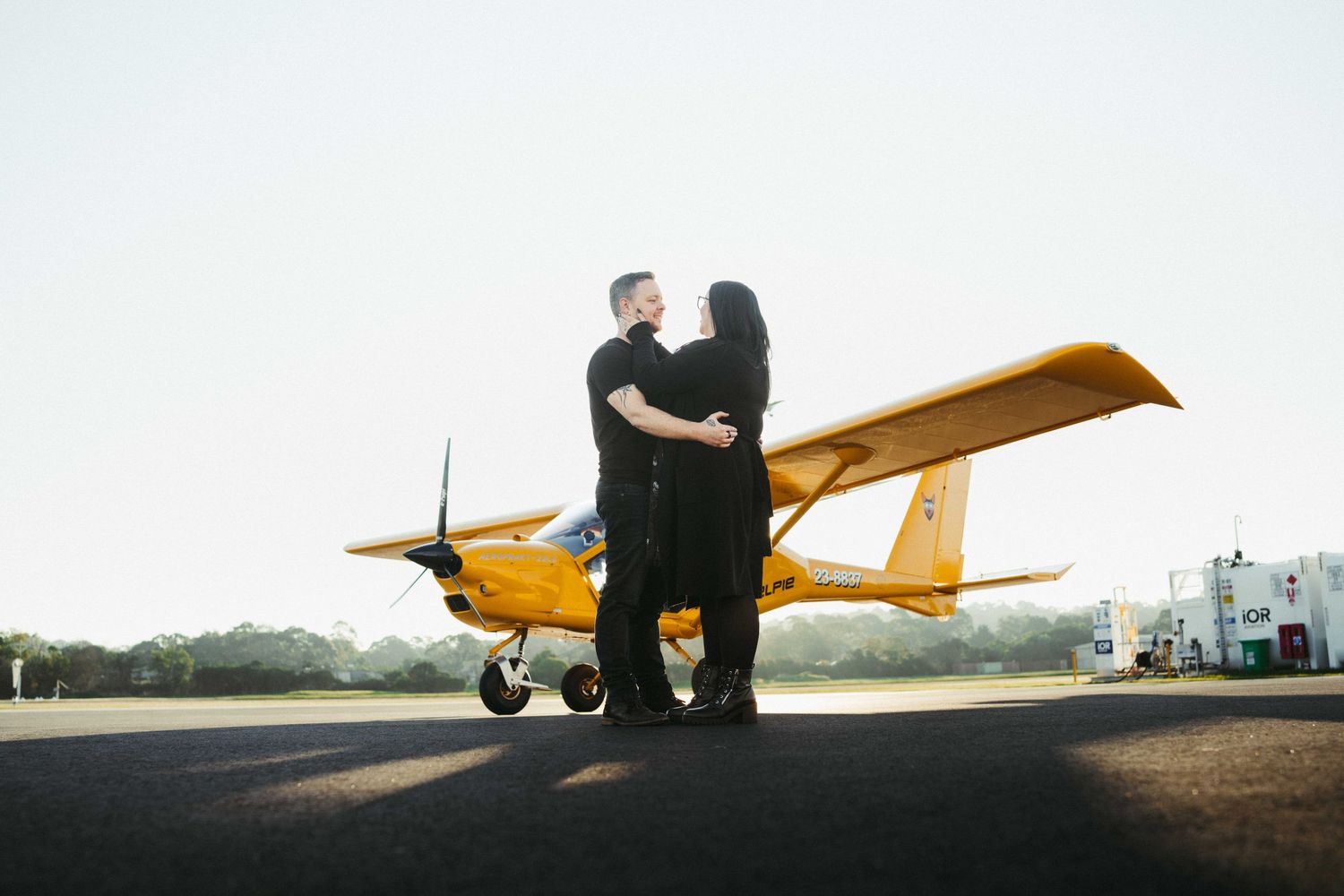 Couple embracing in front of a vibrant yellow small airplane on a sunny airstrip.