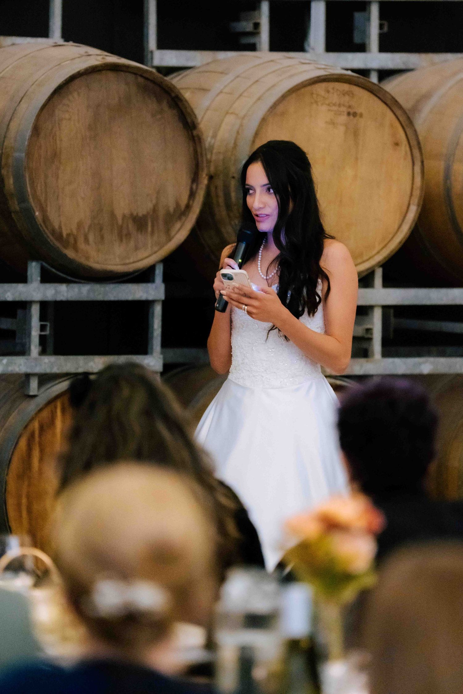 Dark-haired bride in white lace gown gives speech holding microphone in front of wooden wine barrels.