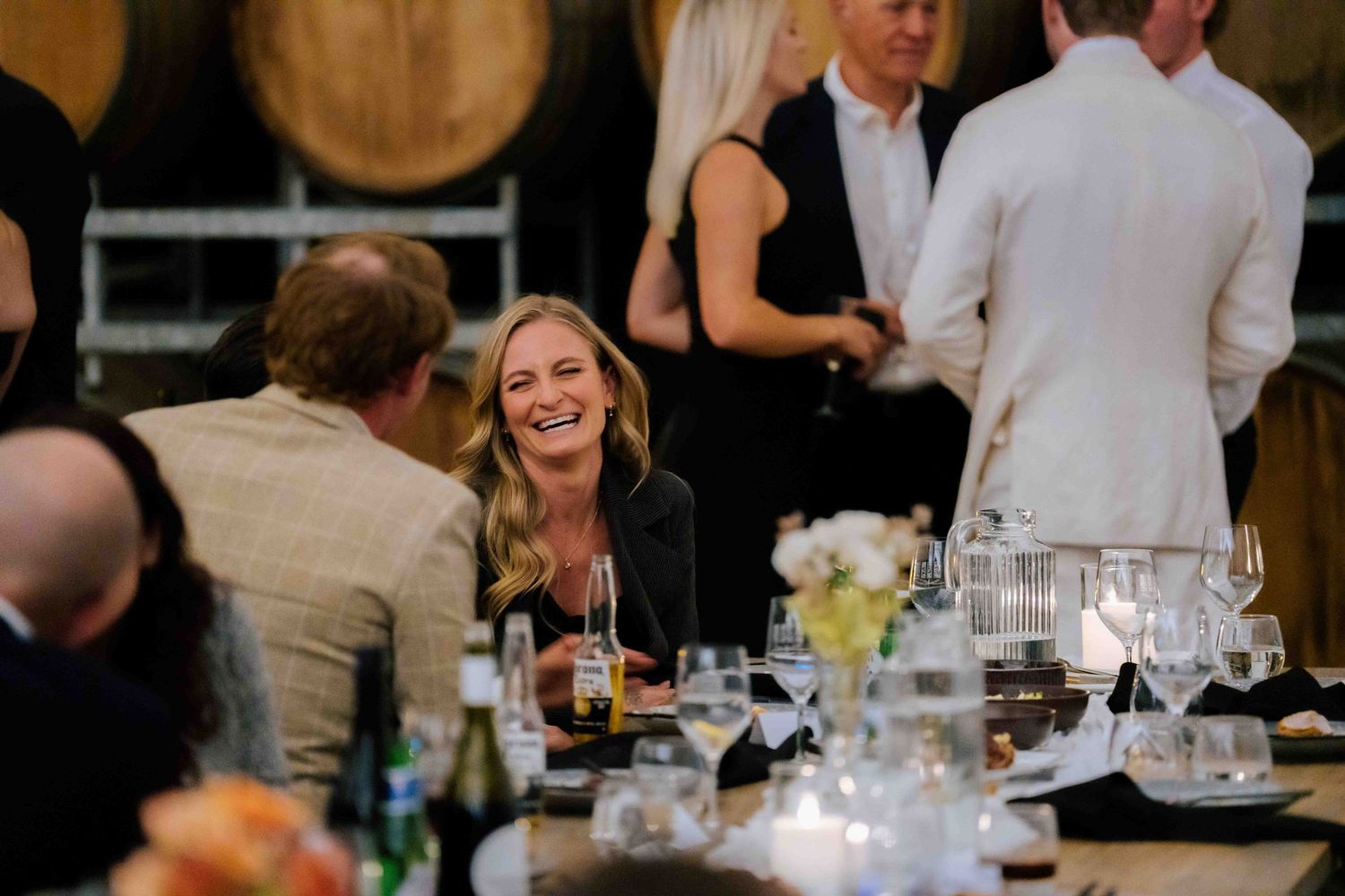 A woman laughs joyfully at an elegant winery dinner reception with wine glasses and floral centerpieces on the table.