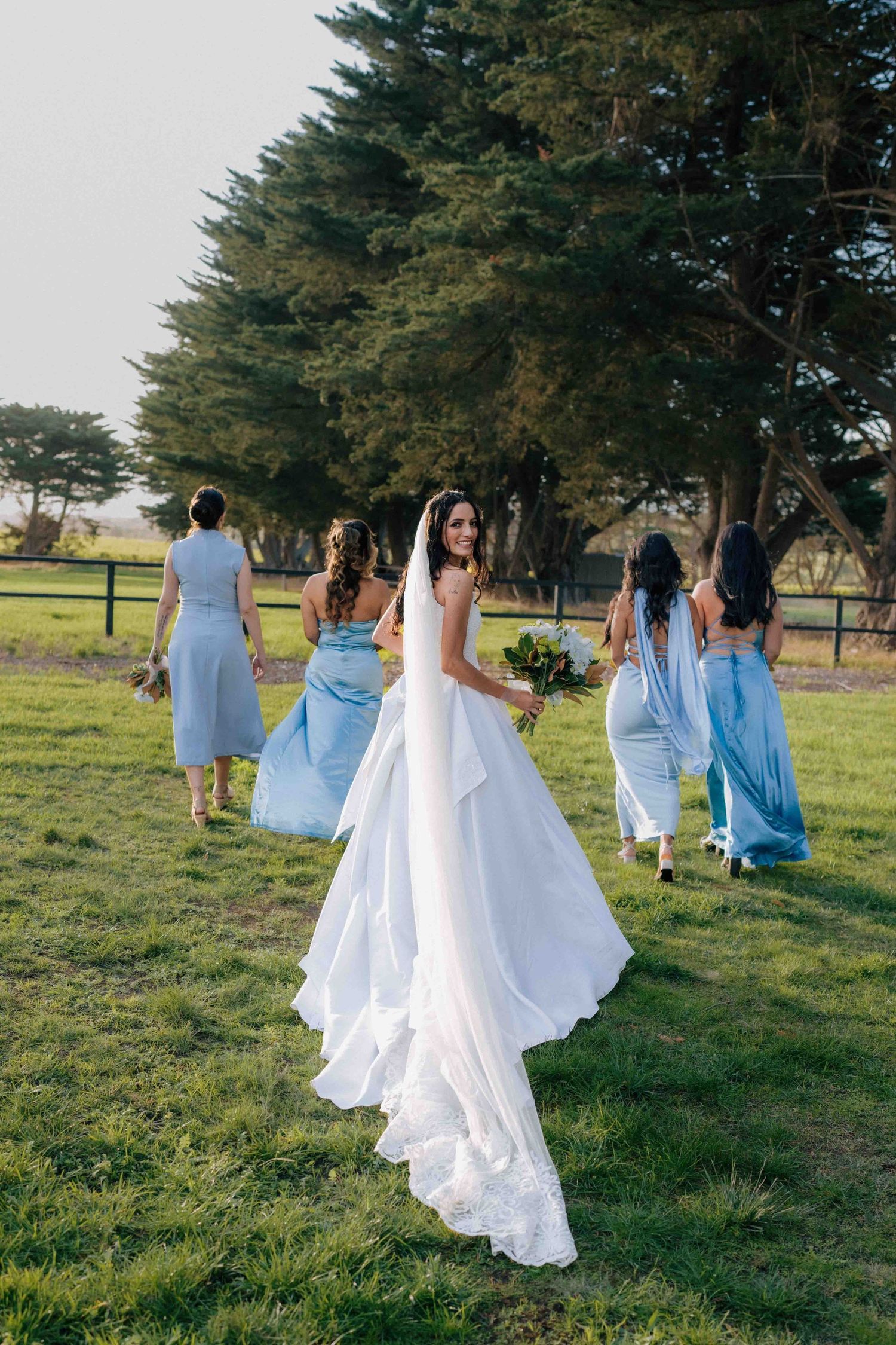 Bride in white gown holds bouquet while bridesmaids in blue dresses walk ahead on green lawn.