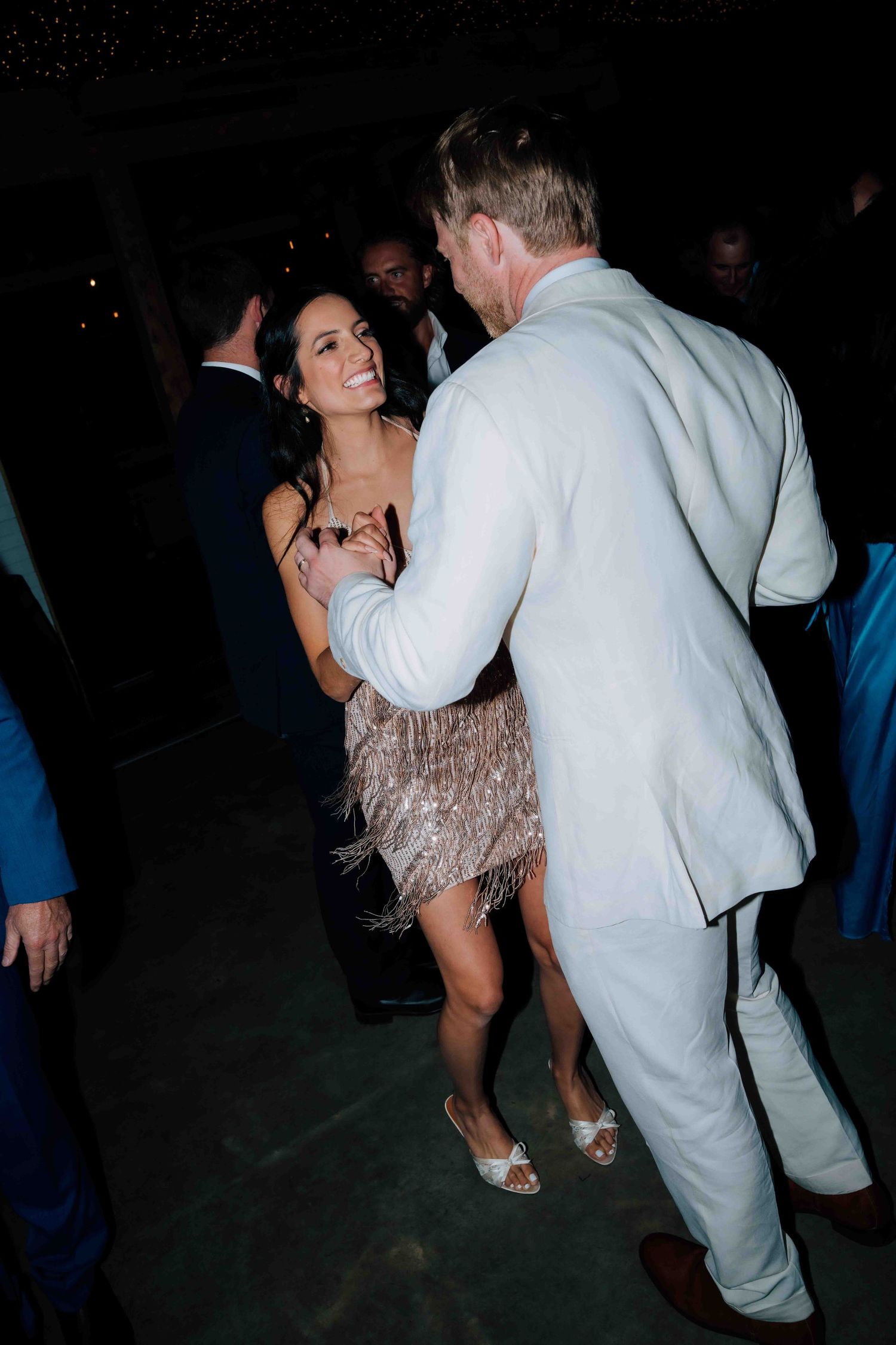 Bride in sparkly fringe dress dancing and laughing with groom in white suit at evening wedding reception.