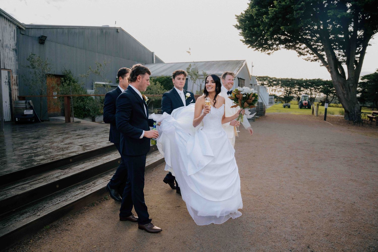Two groomsmen carry the bride in her white gown outside a rustic wedding venue at dusk.