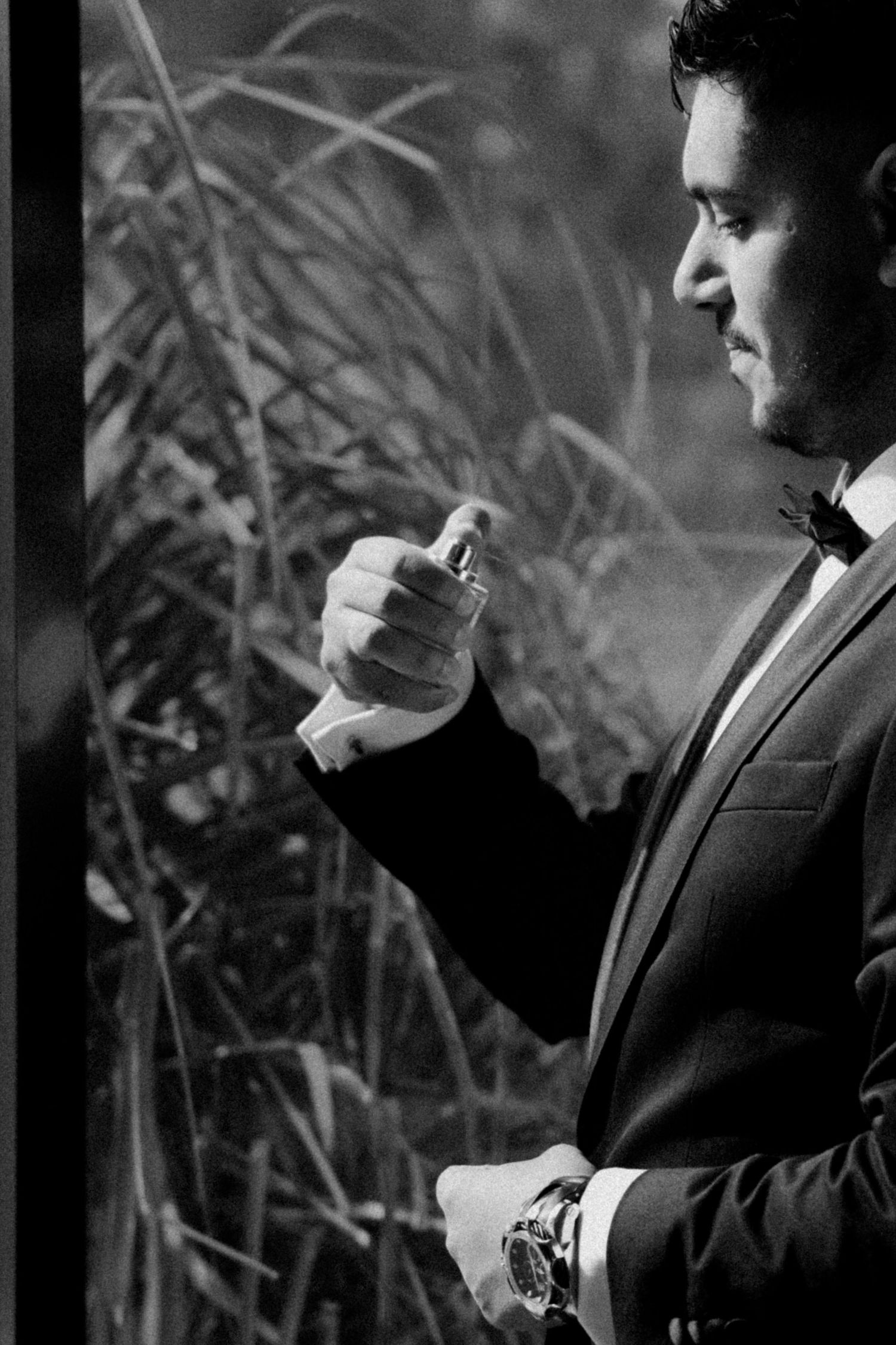 Dapper groom in tuxedo applying cologne outdoors among tall reeds, captured in elegant black and white.