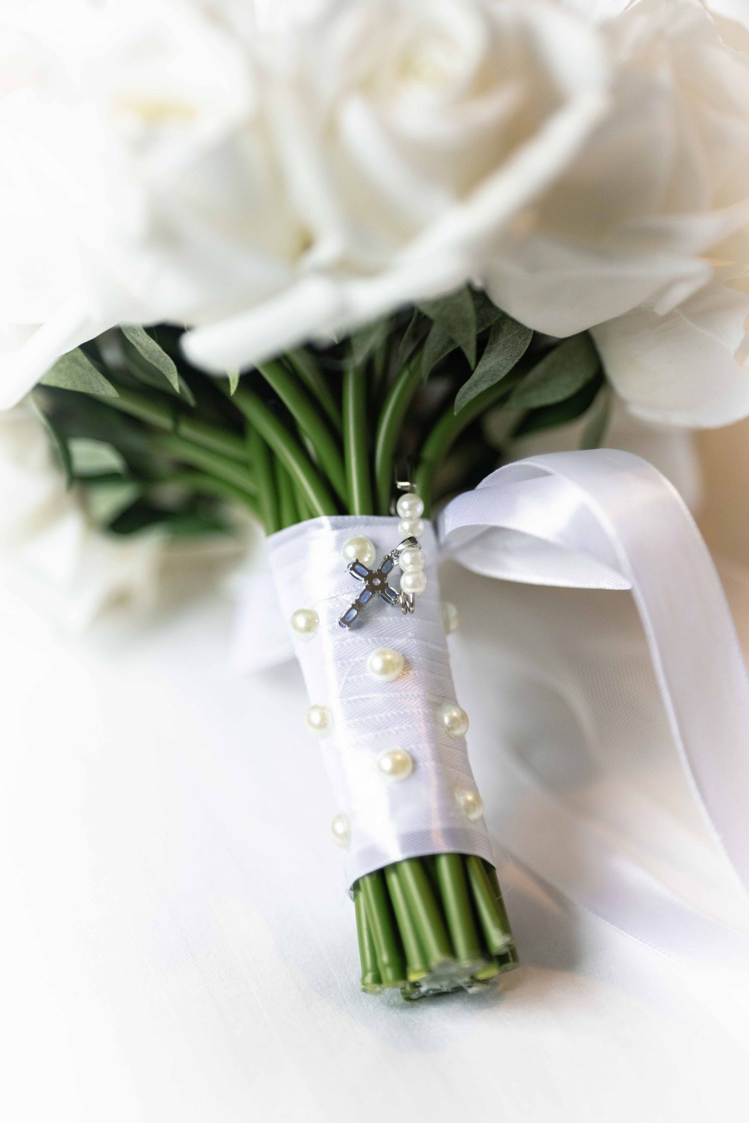 Elegant white rose bridal bouquet wrapped in white ribbon with pearl and cross embellishments.