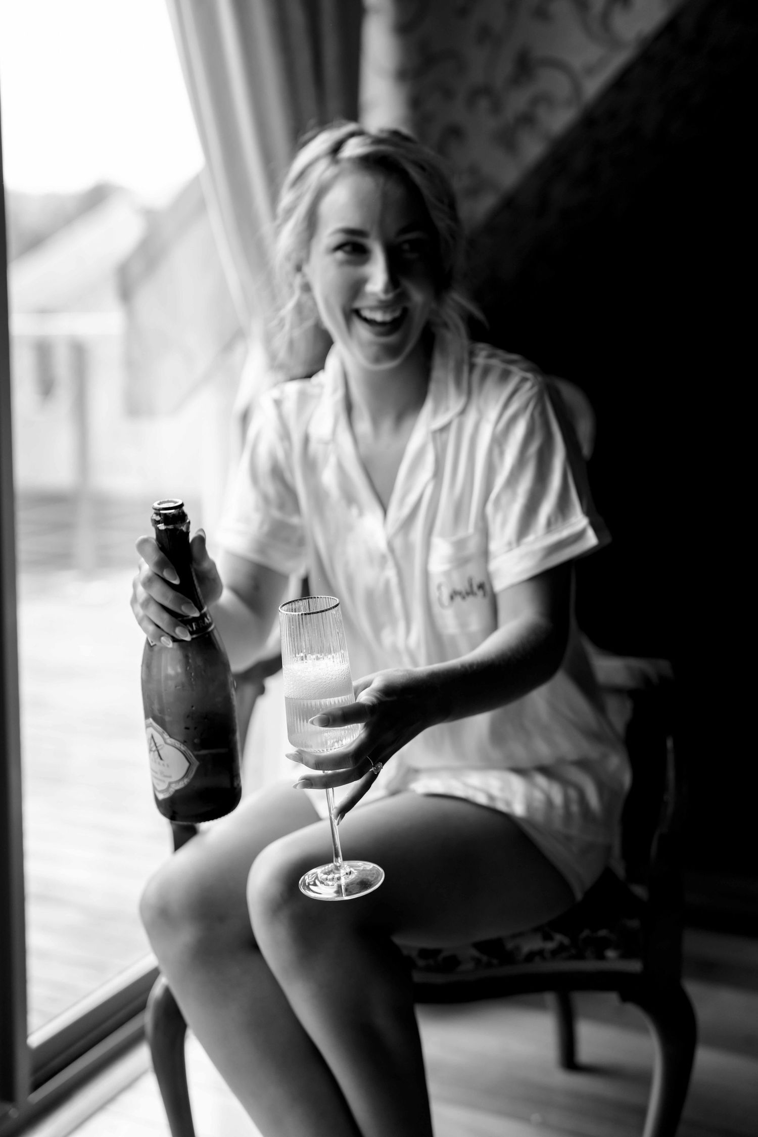 Black and white photo of a smiling blonde bride in pajamas holding champagne bottle and glass by a window.