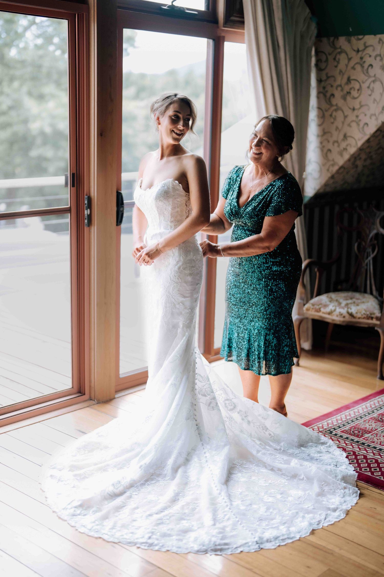 Bride in white lace gown with train stands with older woman in teal dress by window.