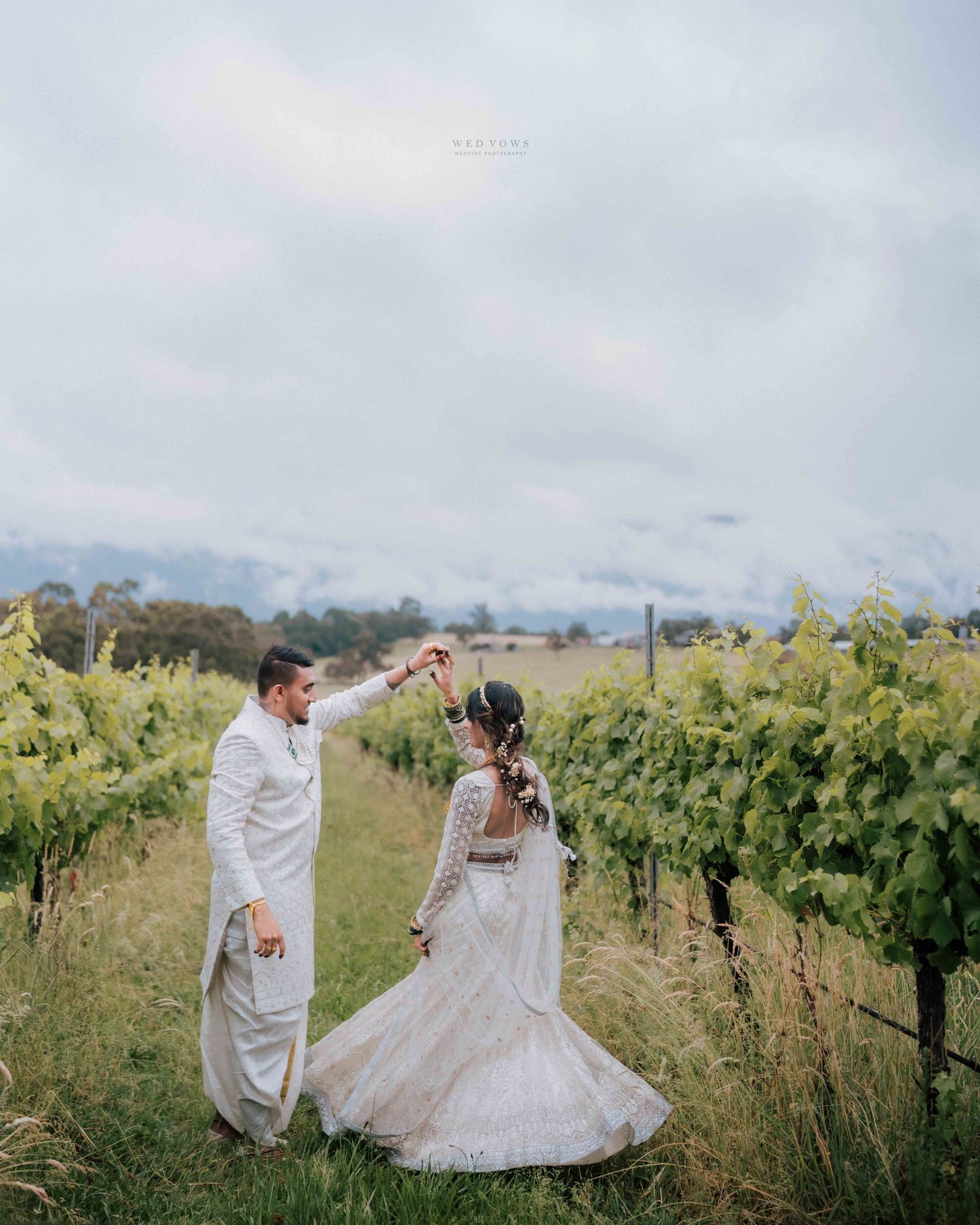 Indian groom twirls his bride in white wedding attire among lush green vineyard rows.
