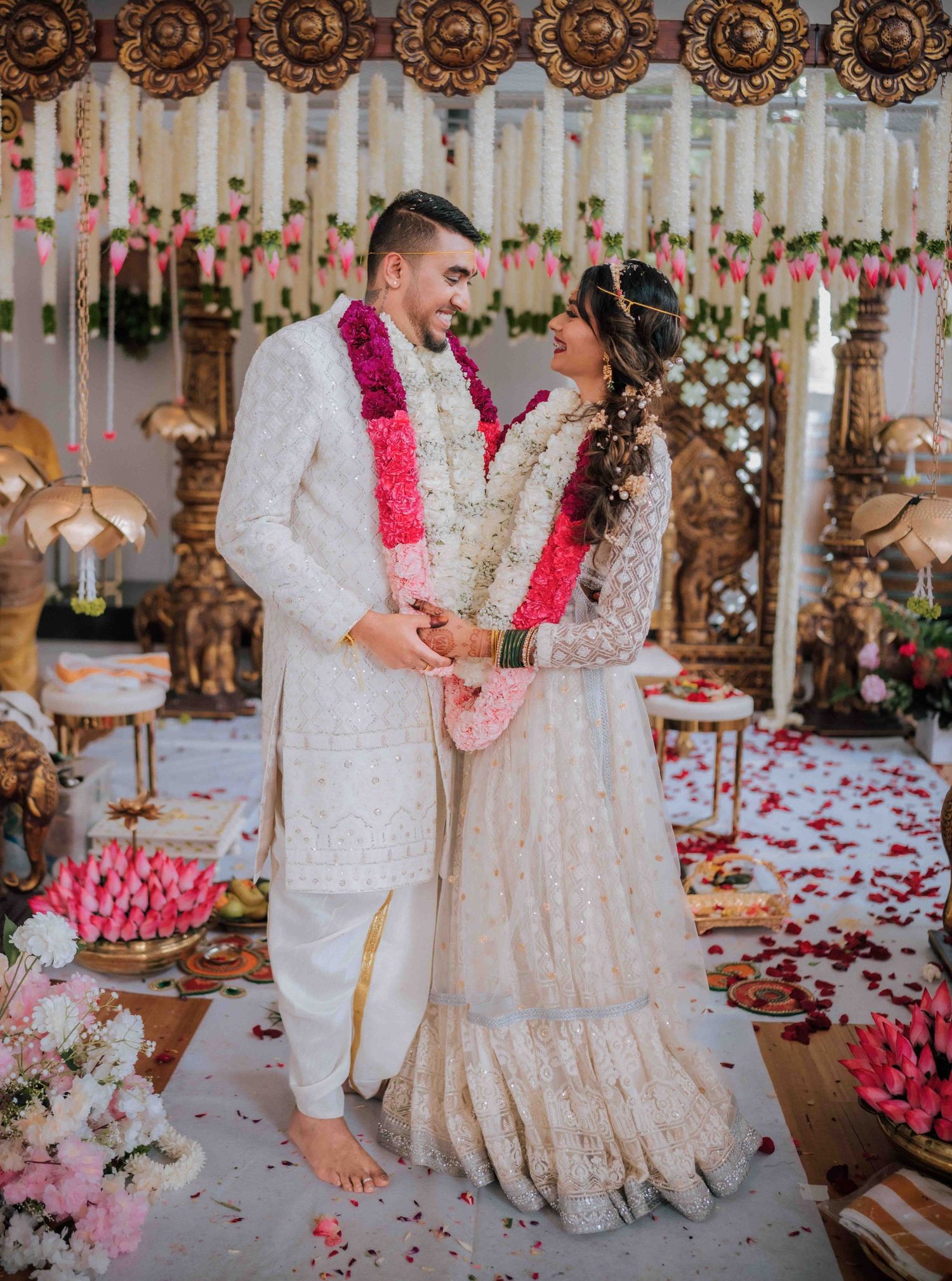 Indian bride and groom in white outfits exchange garlands at an ornate mandap decorated with flowers and candles.