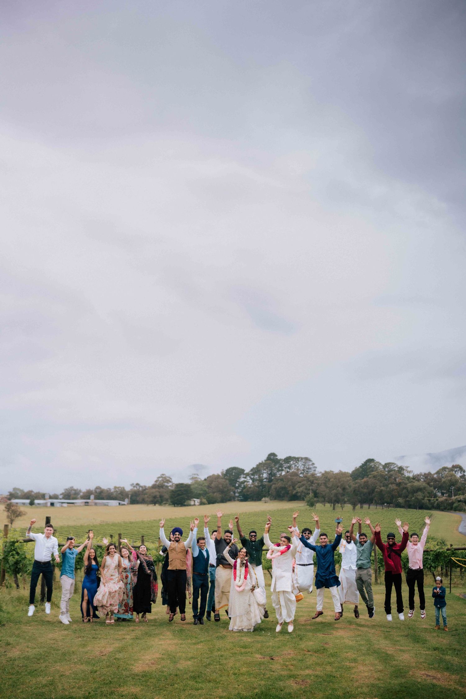 Joyful wedding group photo with guests jumping and raising arms in a lush green vineyard under cloudy skies.