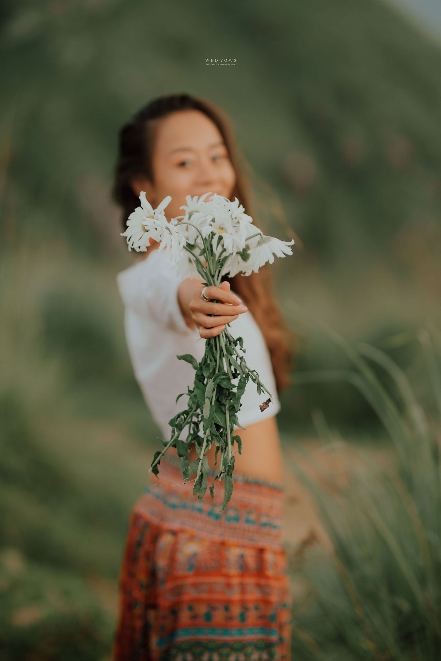 A smiling woman in a white top and tribal skirt holds out a bouquet of white daisies toward the camera outdoors.
