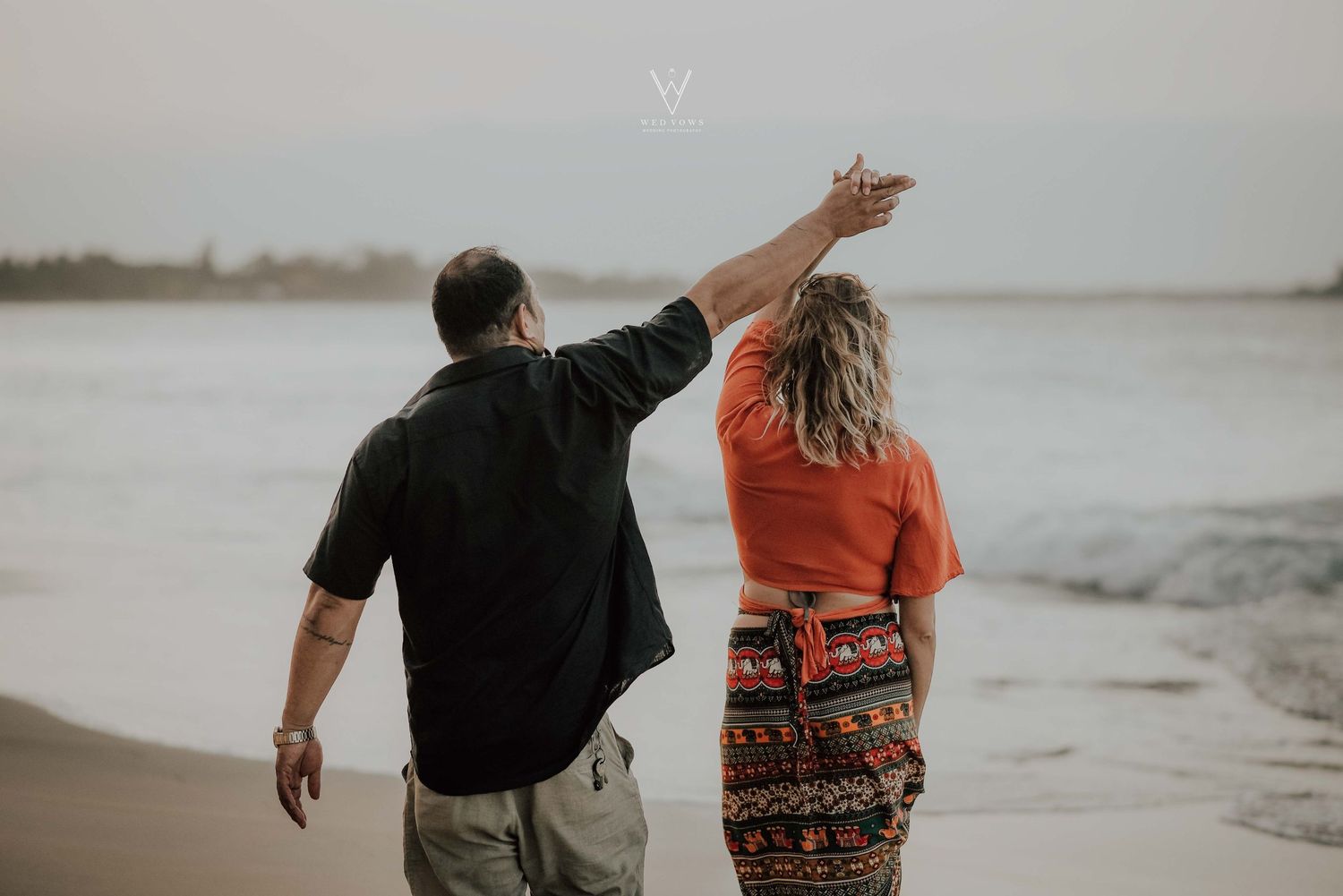 Couple walking on beach from behind, man raising woman's hand while pointing toward the horizon.
