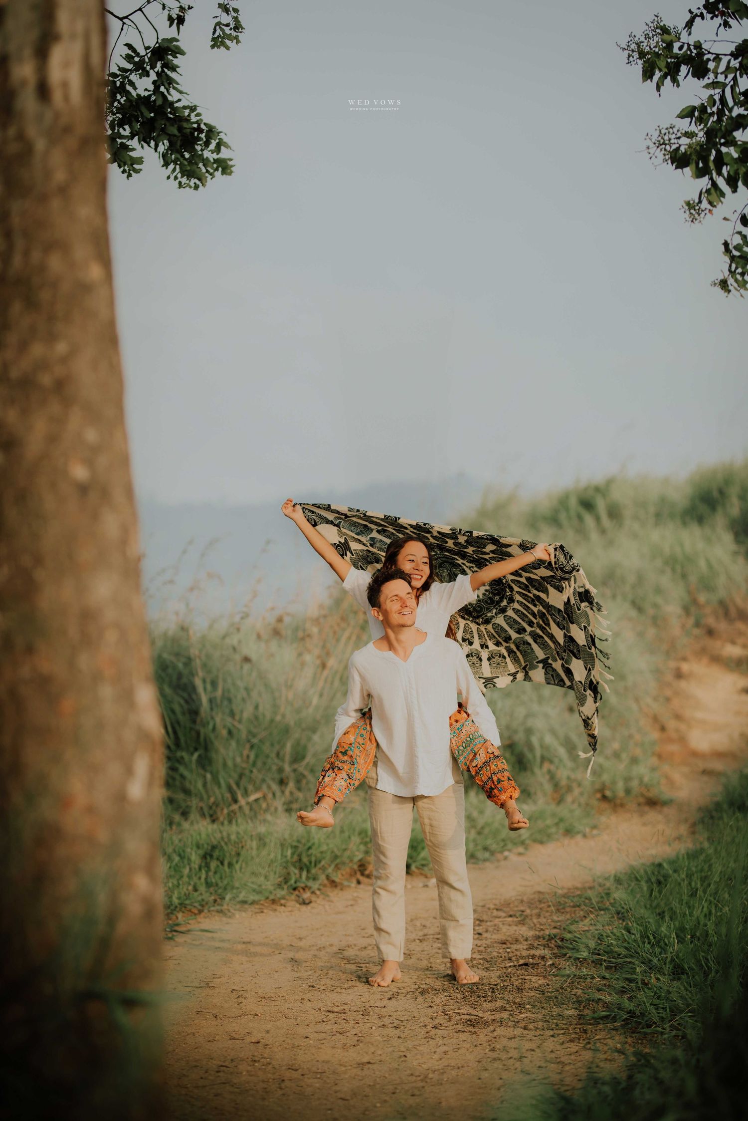 Man carrying woman on shoulders with flowing scarf on scenic nature trail.