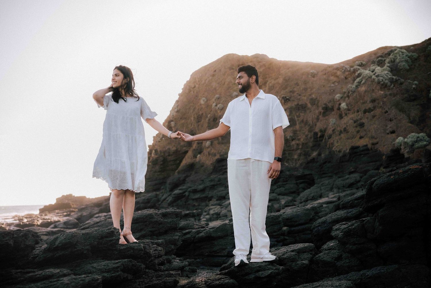 Couple in matching white outfits holding hands on dark volcanic rocks by the sea.