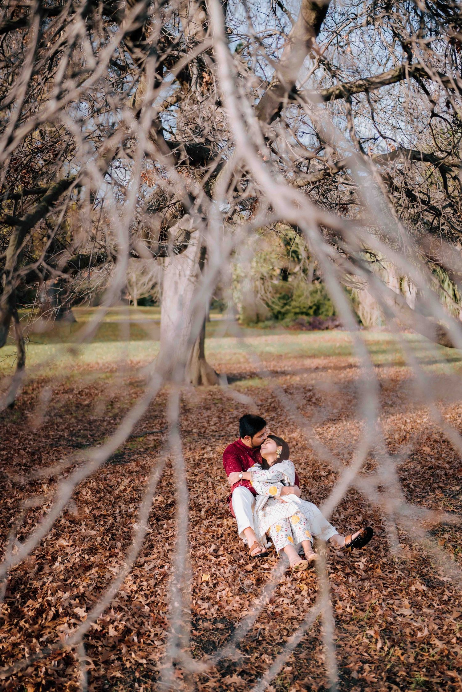 Romantic couple sitting among autumn leaves, framed by delicate bare branches in a sunlit park.