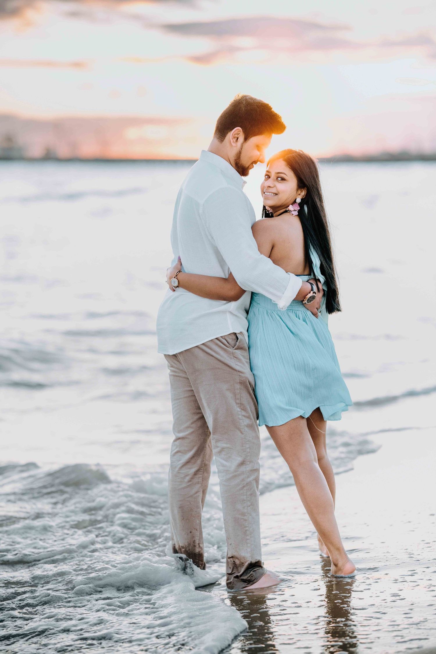 Couple embracing romantically on beach at golden sunset, woman in blue dress smiling over shoulder.