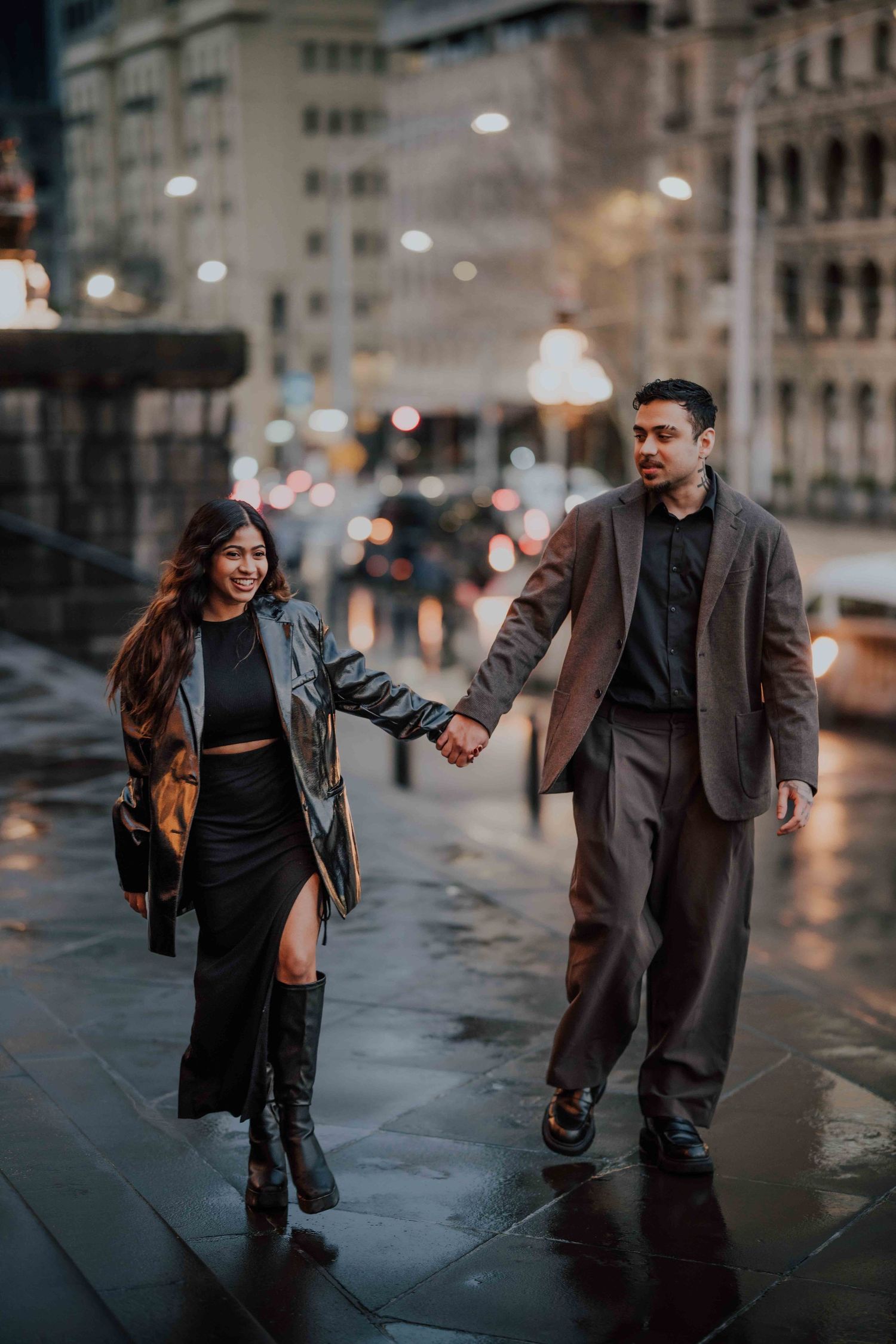 Smiling couple holding hands and walking on a wet city street at night with glowing lights.
