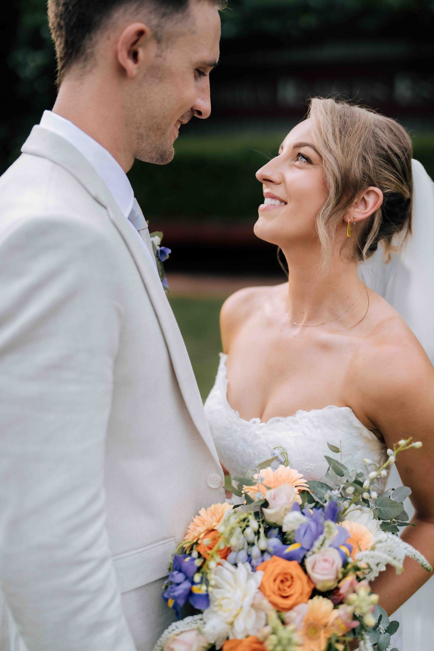 Bride smiles up at groom holding colorful peach and blue bouquet at outdoor wedding.