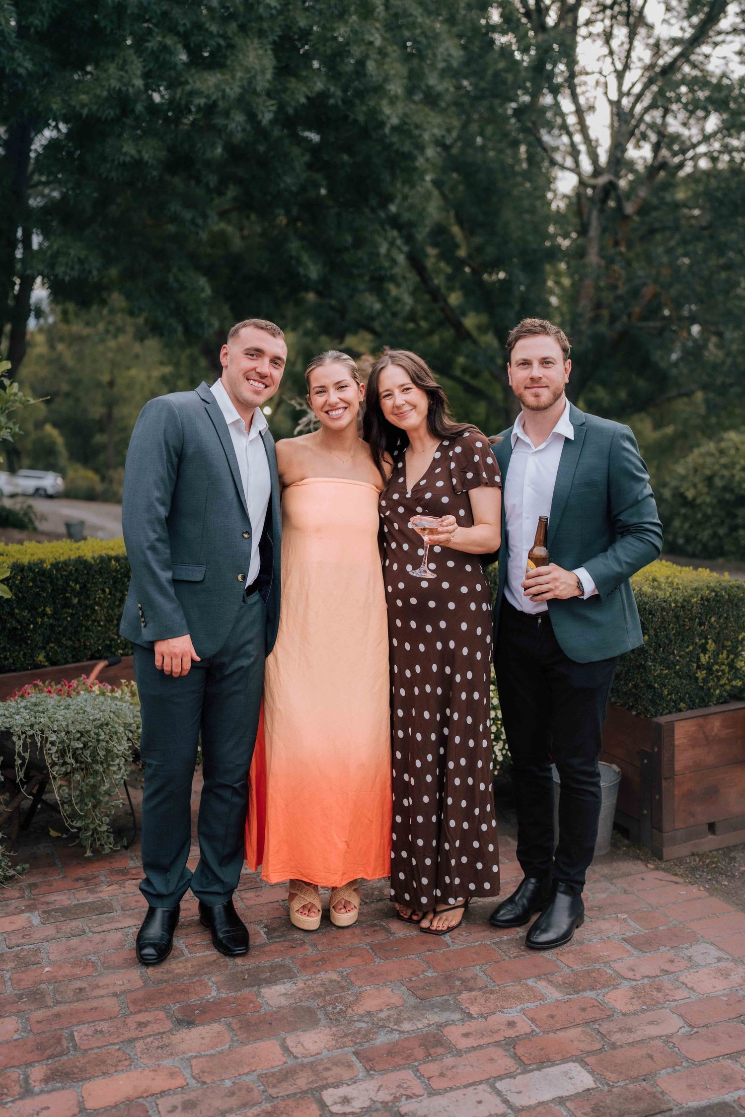 Two couples posing outdoors at wedding reception, wearing formal attire, smiling in front of green hedges.