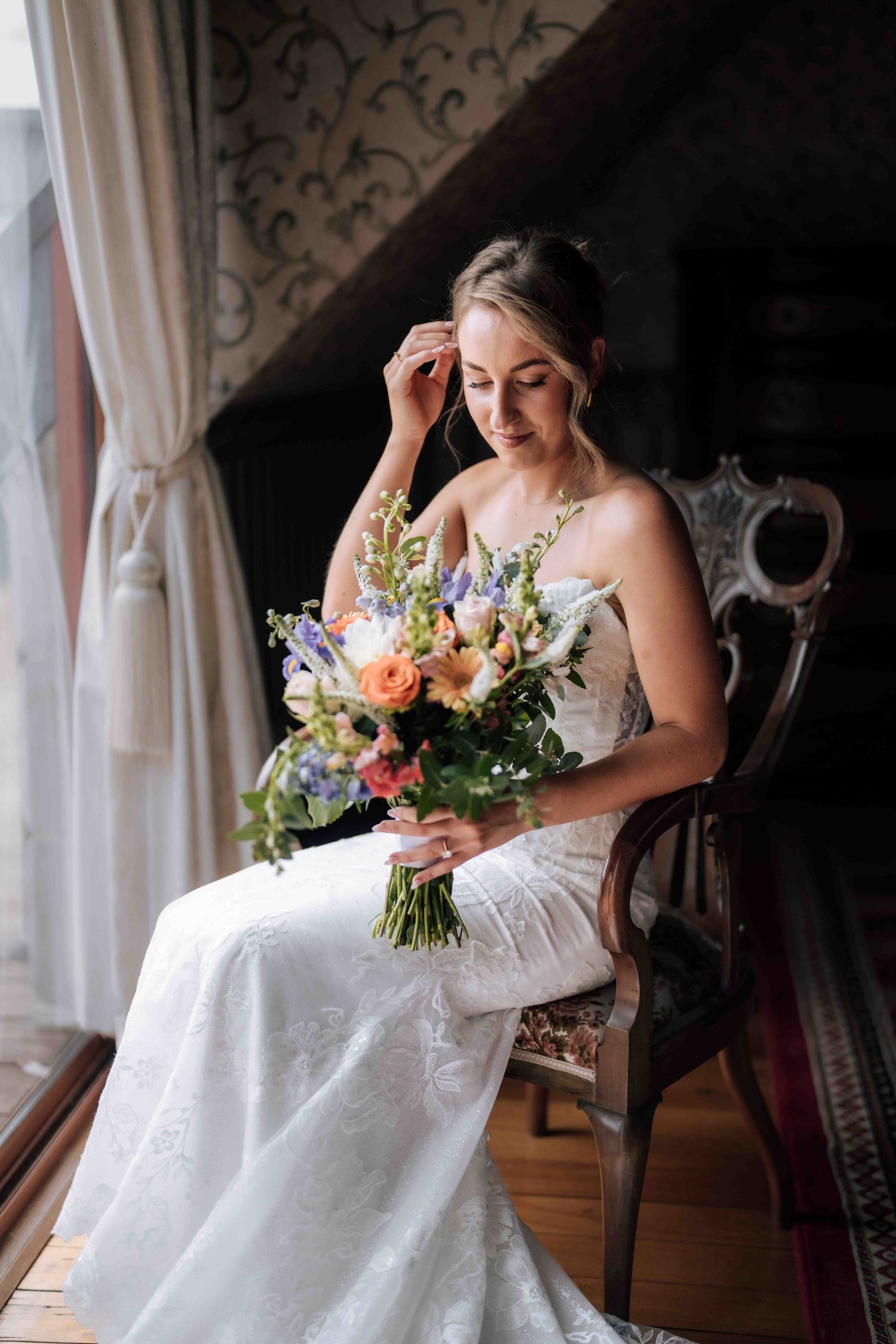 An elegant bride in a white lace gown sits in an antique chair holding a colorful bridal bouquet by a window.