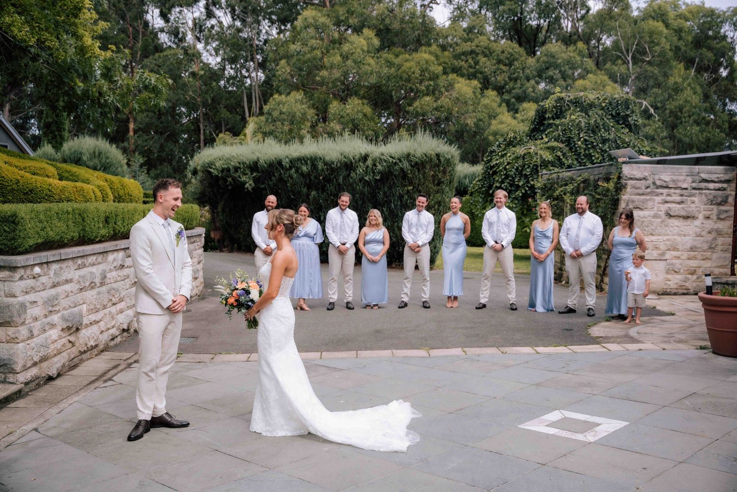 Bride and groom share intimate first look moment on stone terrace with wedding party in background.