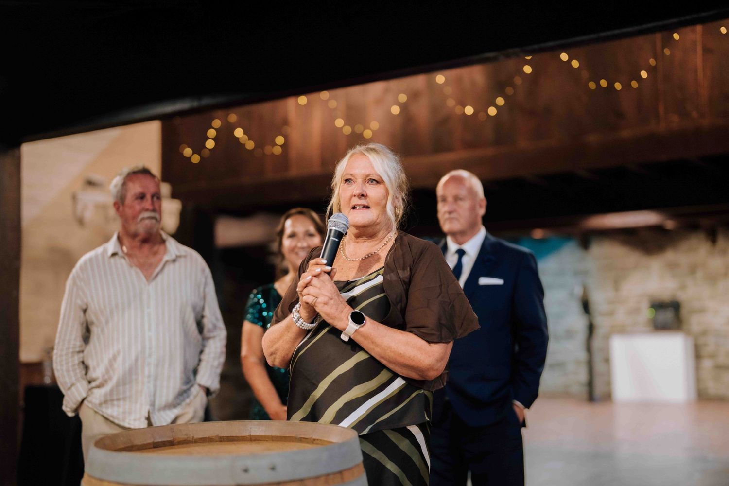 Woman speaking into microphone at wedding reception podium with guests standing behind her.