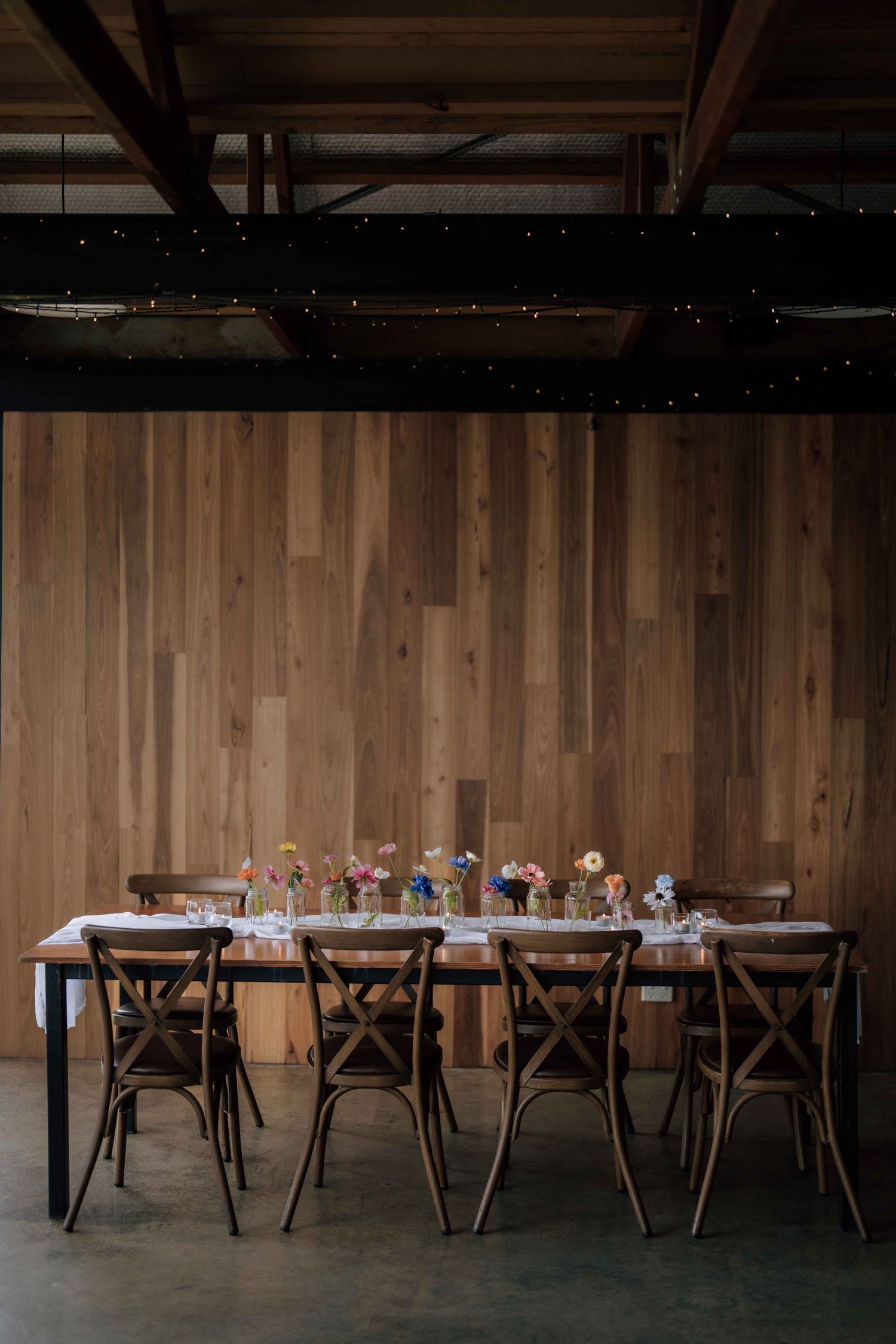 Rustic wedding reception table set for ten guests against warm wood panel wall with fairy lights overhead.