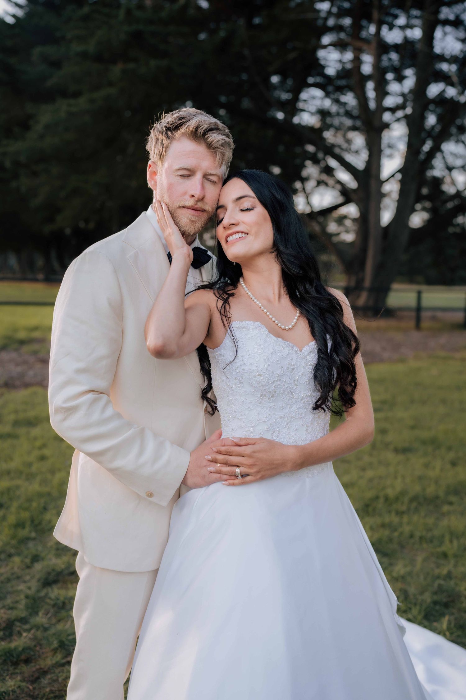 Bride and groom embrace outdoors, she in white gown with pearls, he in cream suit, eyes closed, trees in background.